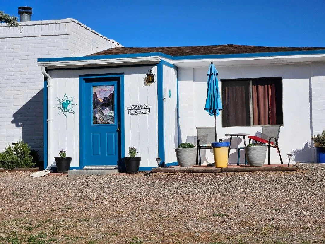 Exterior of a white house with a blue door and trim, a small porch with chairs and an umbrella, potted plants, and a large window with curtains, under a clear blue sky.