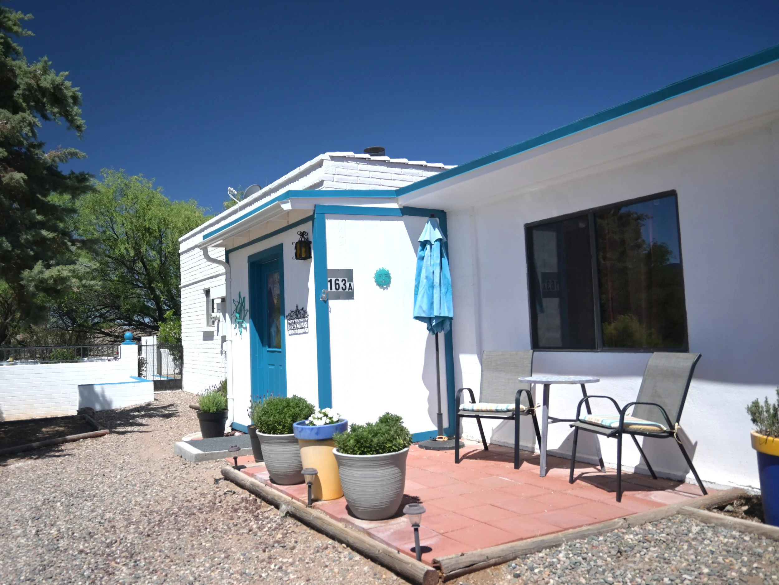 White house with blue trim, outdoor patio with chairs, potted plants, and umbrellas, surrounded by trees and a gravel ground under a clear blue sky.