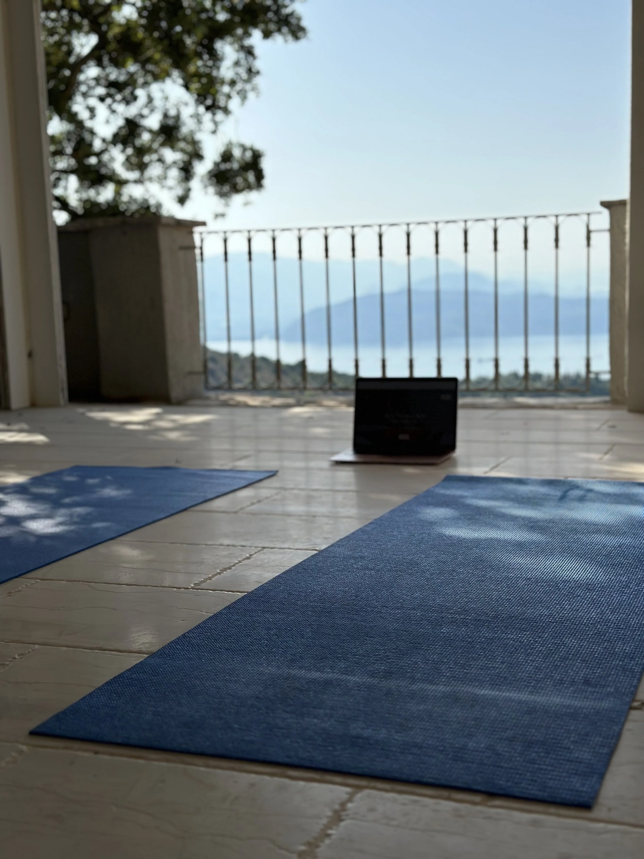 Yoga mats on a wooden floor in front of a balcony with a view of mountains and water and sea in Corfu Greece.