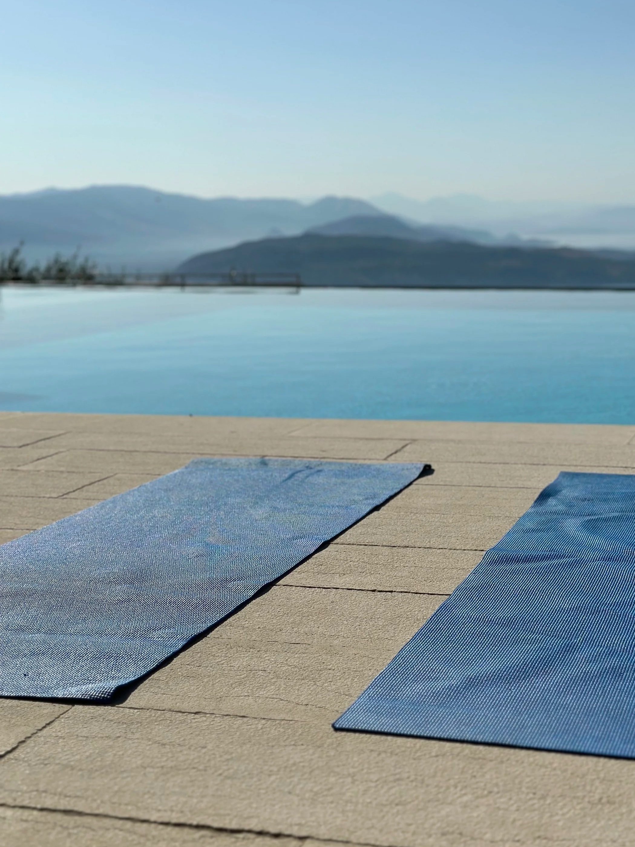 Multiple blue yoga mats placed on a wooden deck near a swimming pool with a mountain range in Corfu, Greece in the background.