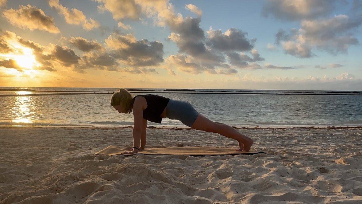 Person practicing yoga on the beach during sunset, in a plank position on a yoga mat.