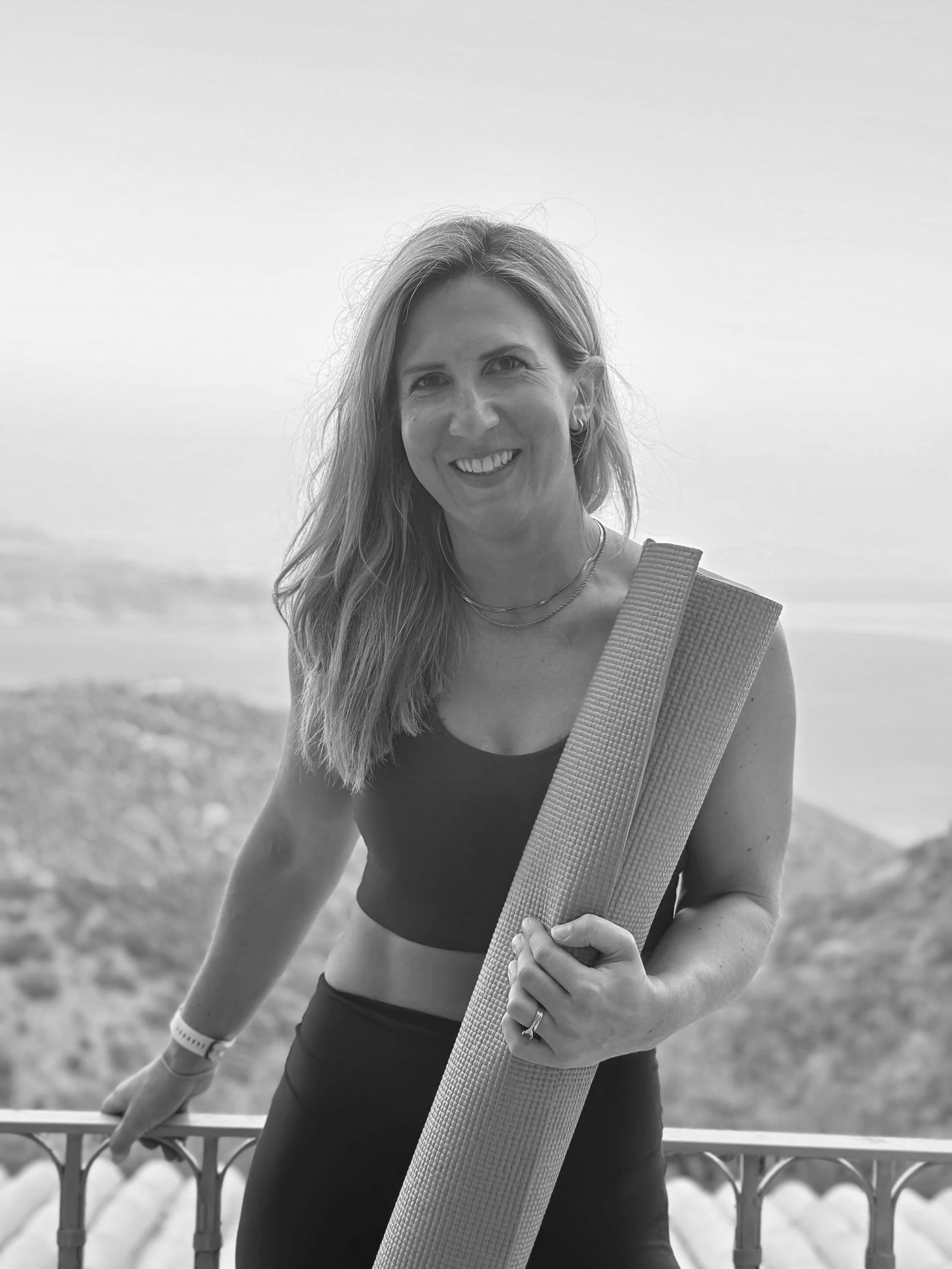 A Pilates instructor woman with long hair smiling, holding a yoga mat, wearing workout clothes, standing on a balcony with railing with view of coastline in Corfu, Greece