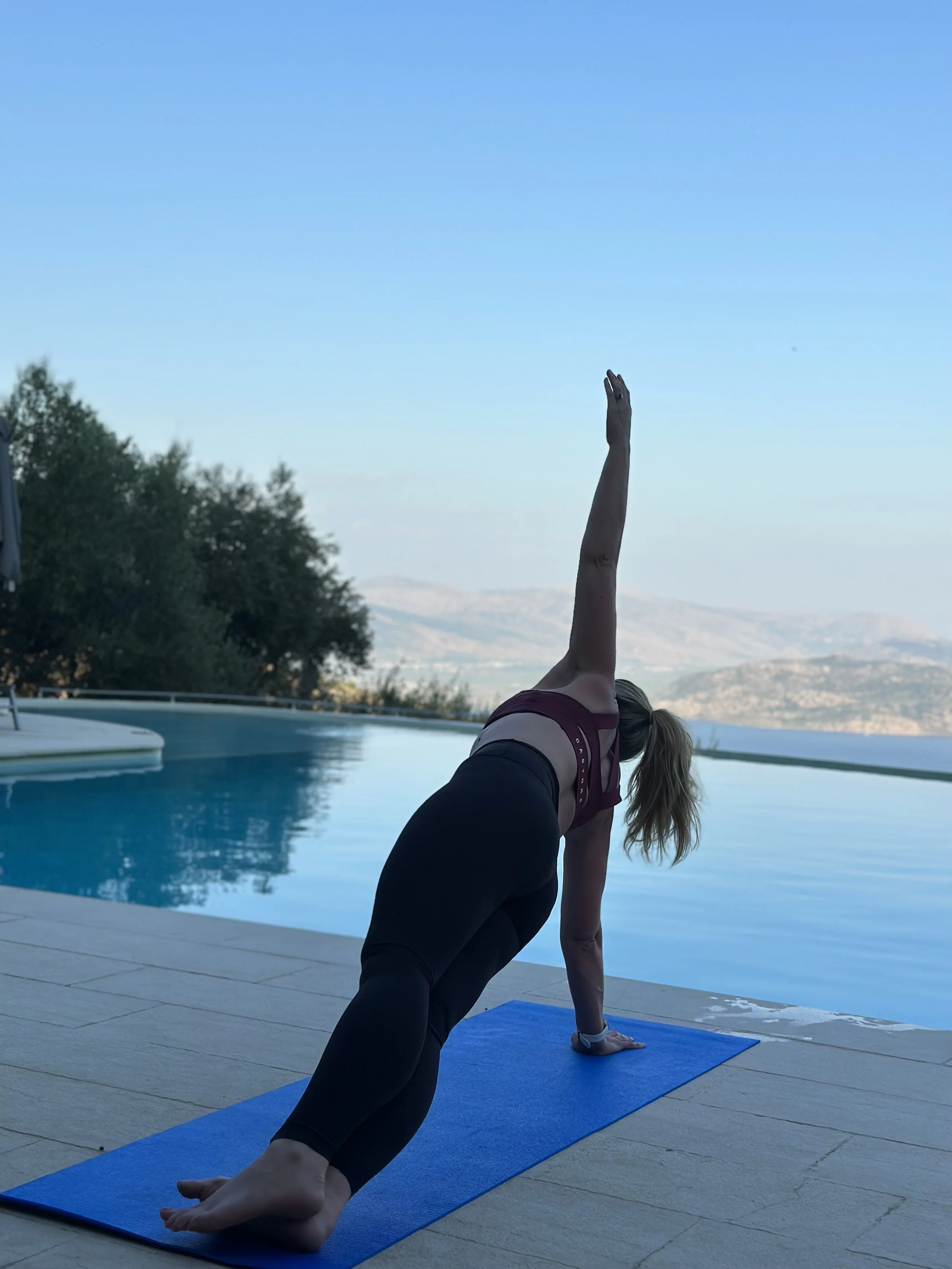 A person practicing yoga outdoors on a blue mat near a swimming pool, performing a side plank pose with an extended arm and one leg lifted, with scenic mountains and trees in the background under a clear blue sky.