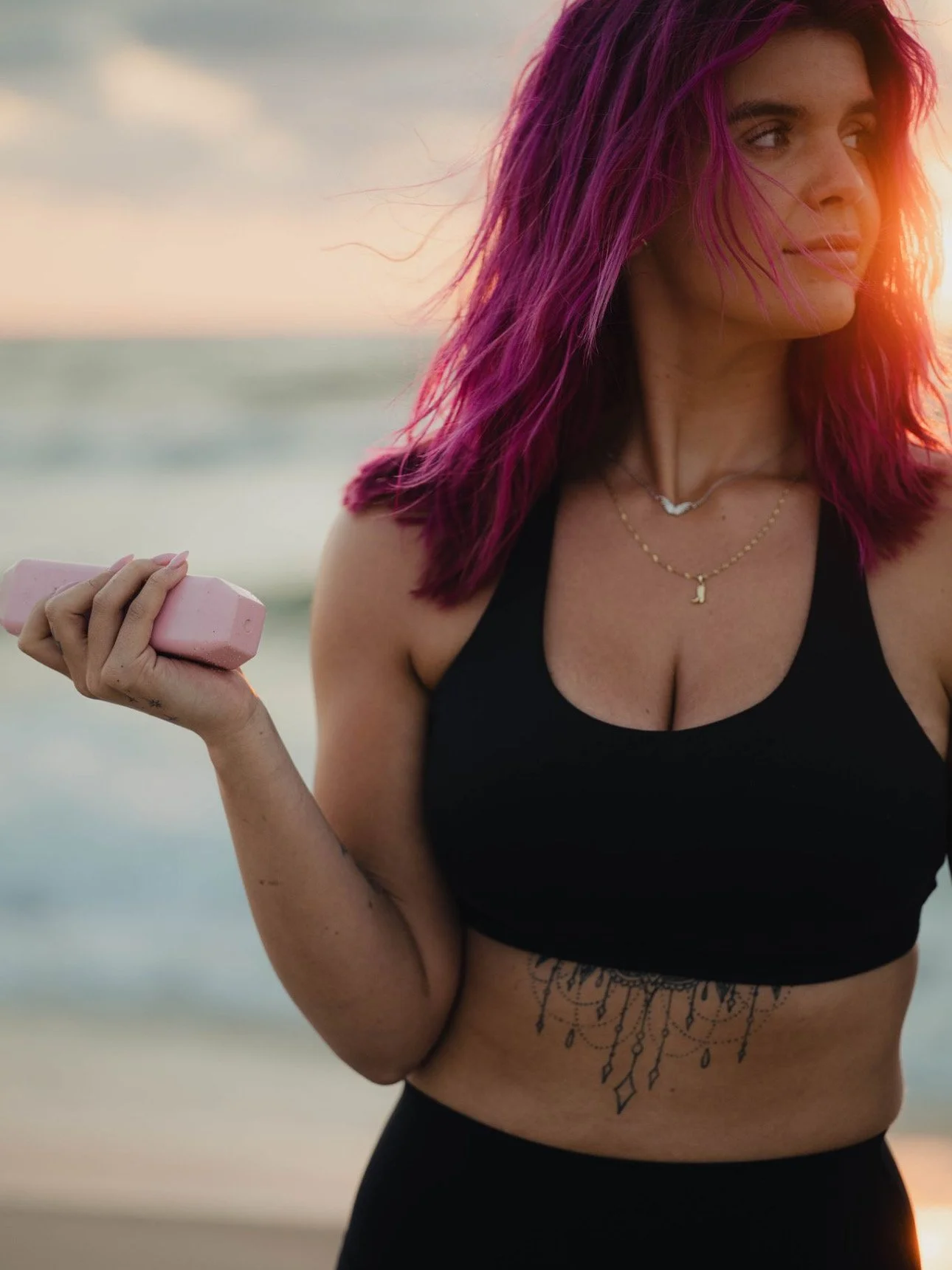 Woman with pink hair holding a pink dumbbell at the beach during sunset, wearing a black sports bra and layered necklaces.