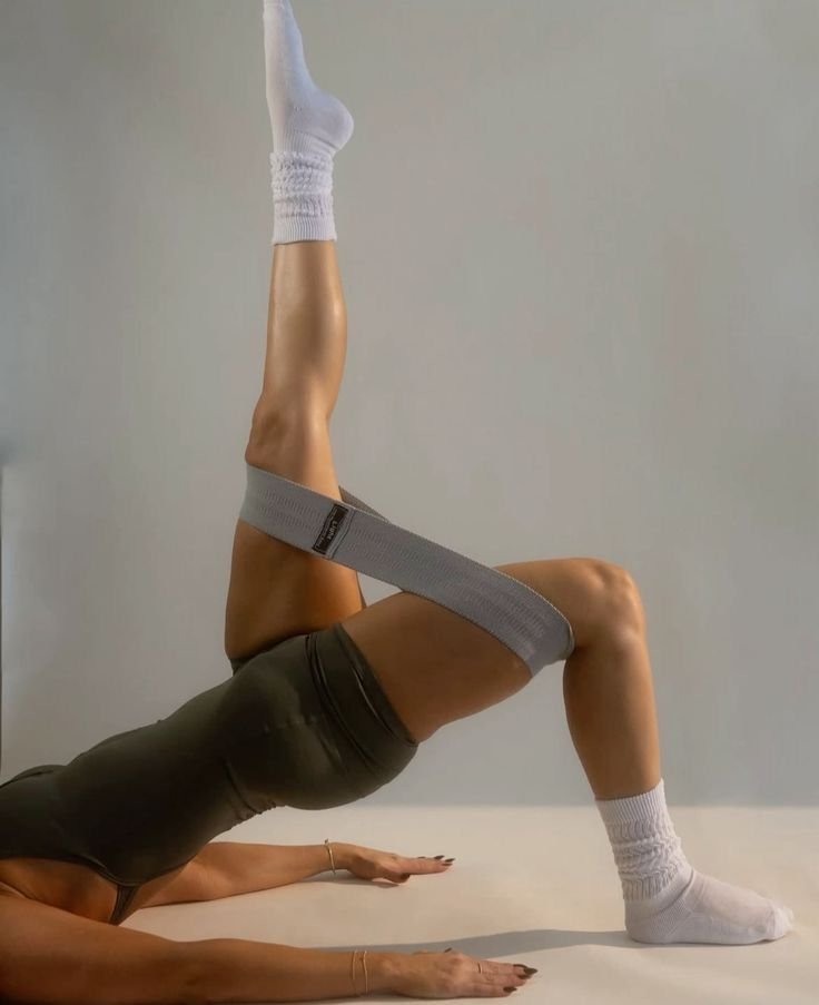 Person practicing yoga on the floor, wearing black athletic attire, white socks, and a gray strap around thighs, performing a bridge pose with one leg extended upward.