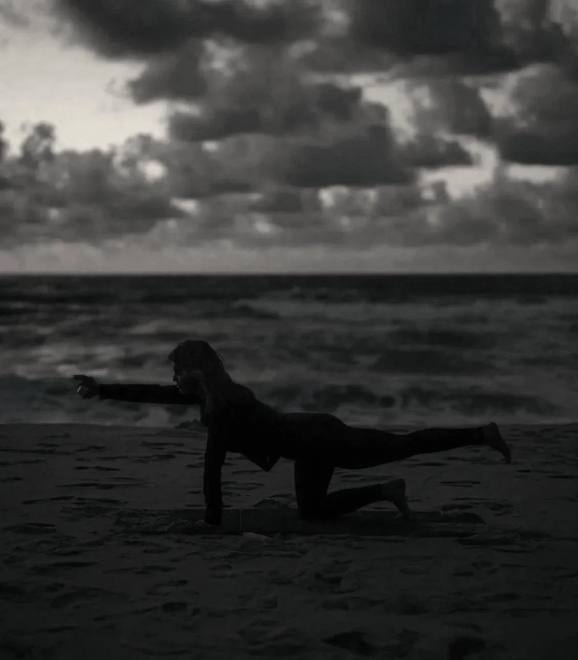 A person practicing yoga or stretching on the beach at sunset or dusk, with the ocean and cloudy sky in the background.