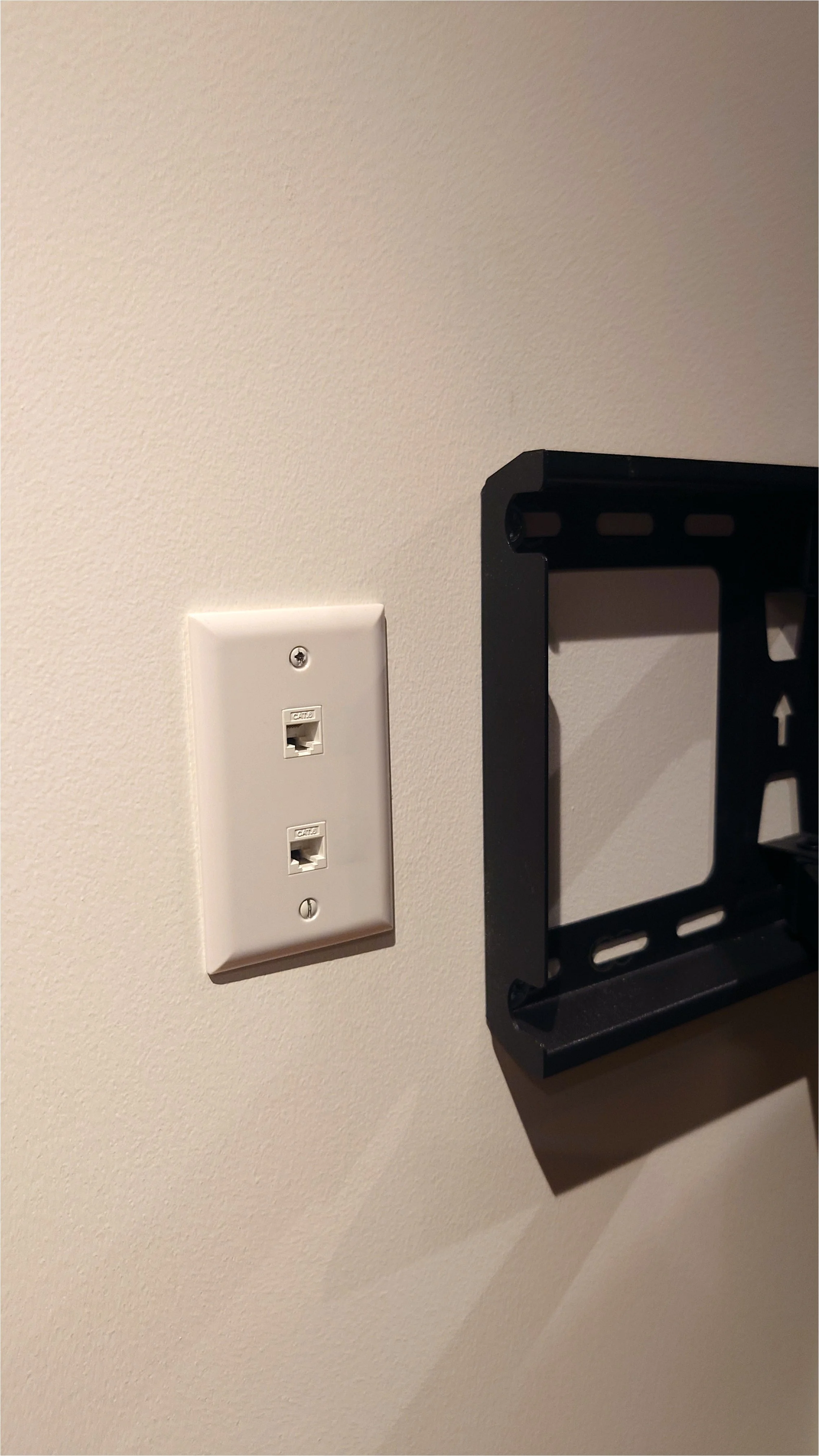 A wall with a white Ethernet wall plate with two ports, and a black metal TV mount next to it.