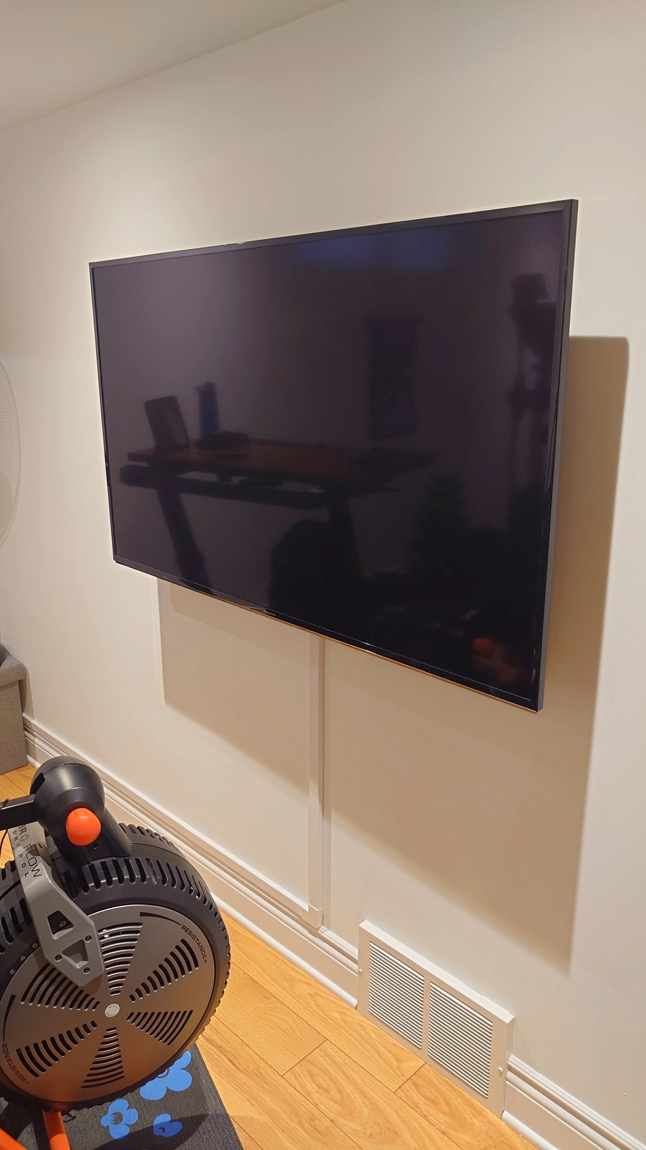 A large flat-screen TV mounted on a beige wall in a room. Below the TV, there is a white vent and a section of a white baseboard.