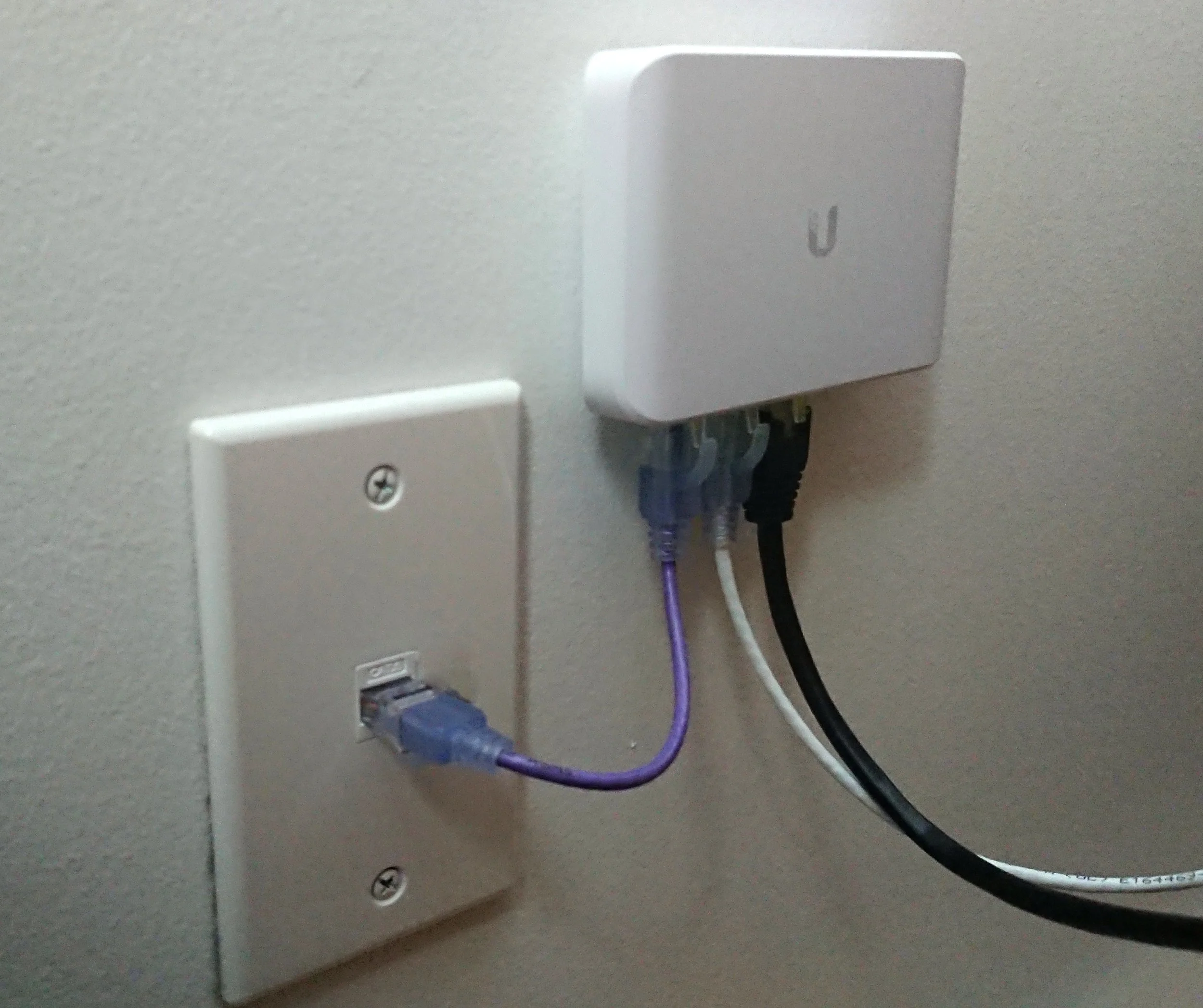 Close-up of a wall socket with a white cover plate, with multiple cables plugged in and connected to a UniFi network switch.