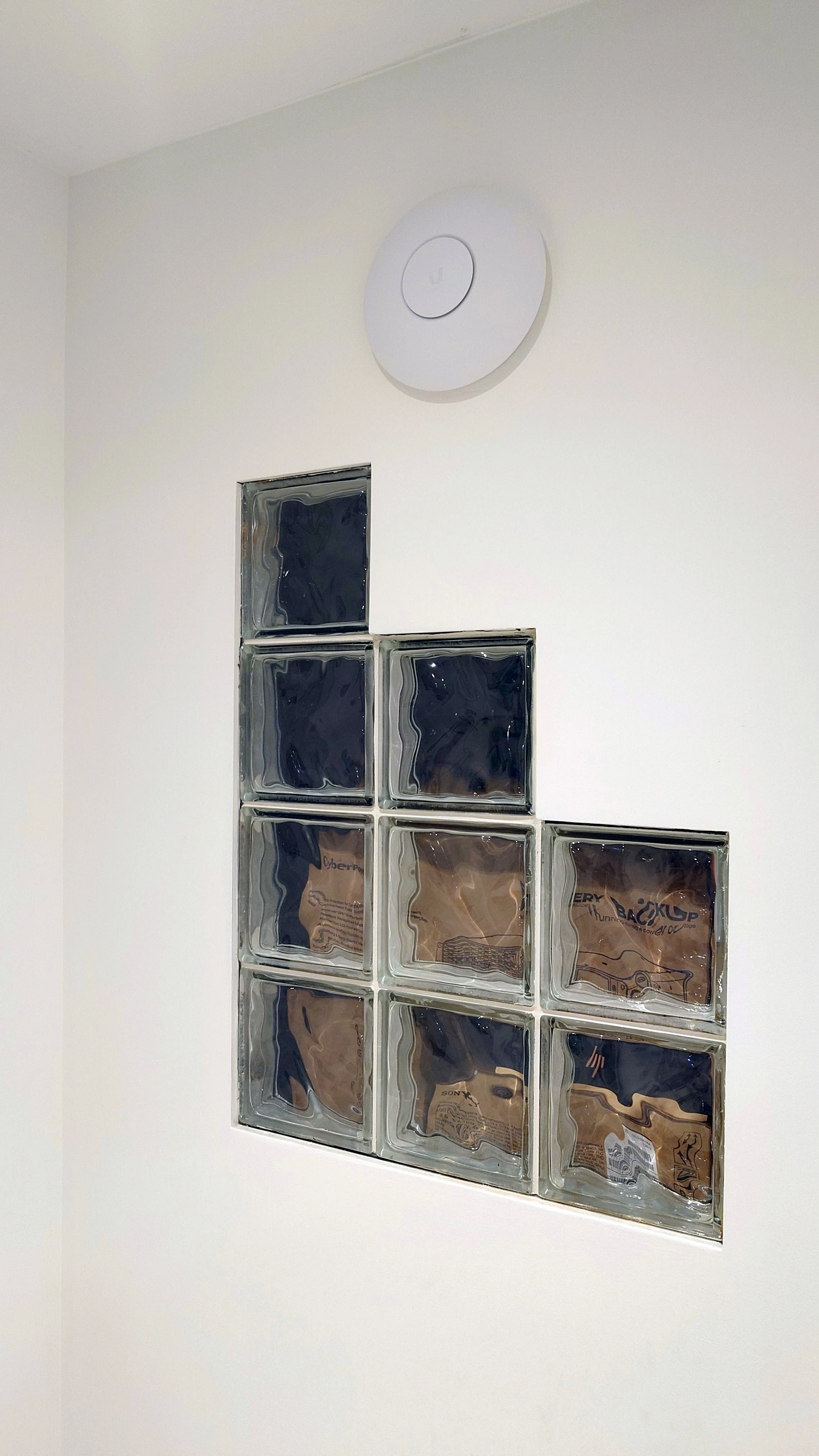 White wall with a circular UniFi wireless access point and a rectangular glass block window.