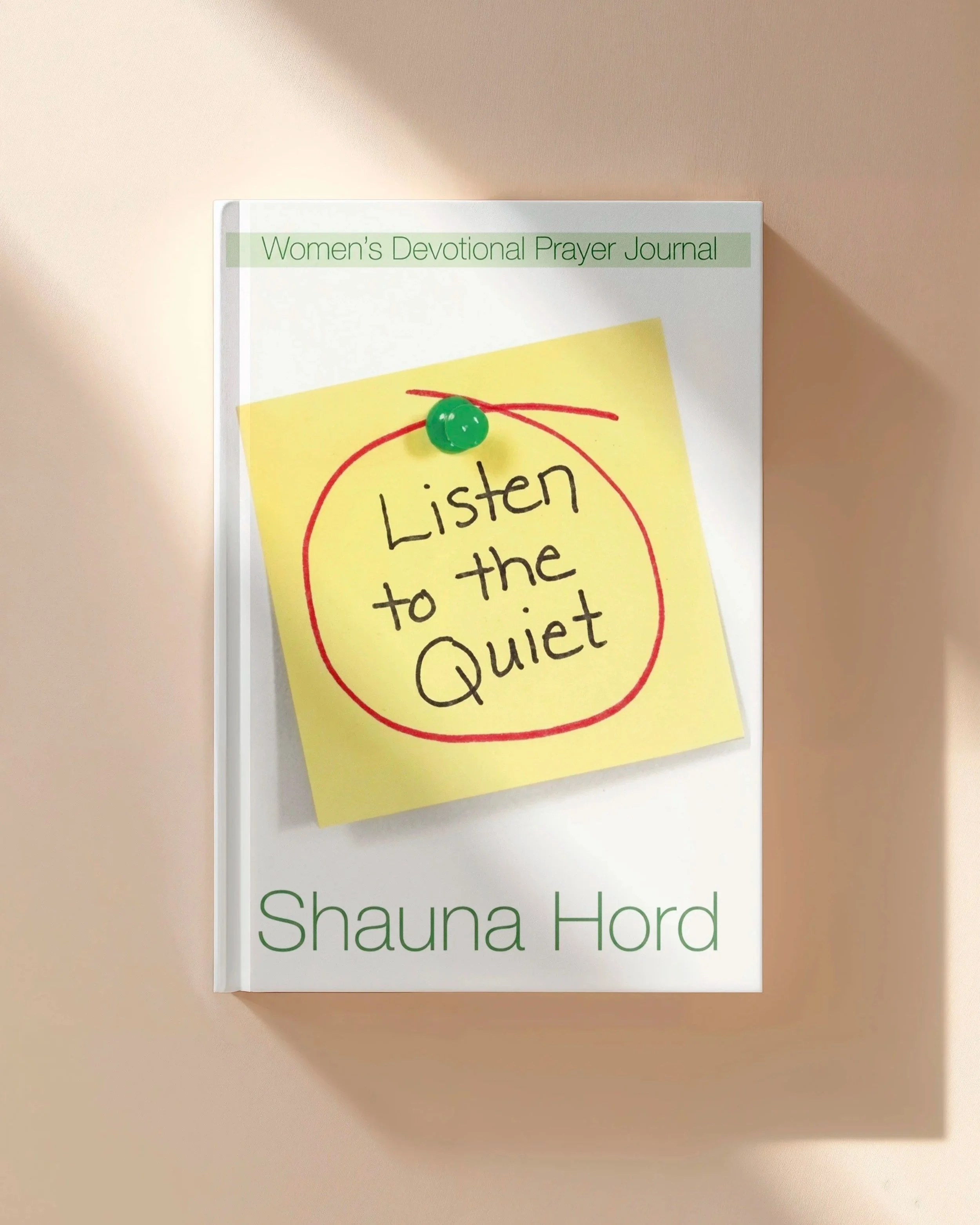 A book titled "Women's Devotional Prayer Journal" by Shauna Ford is attached to a beige wall with a yellow sticky note that says "Listen to the Quiet" and is pinned with a green pushpin.