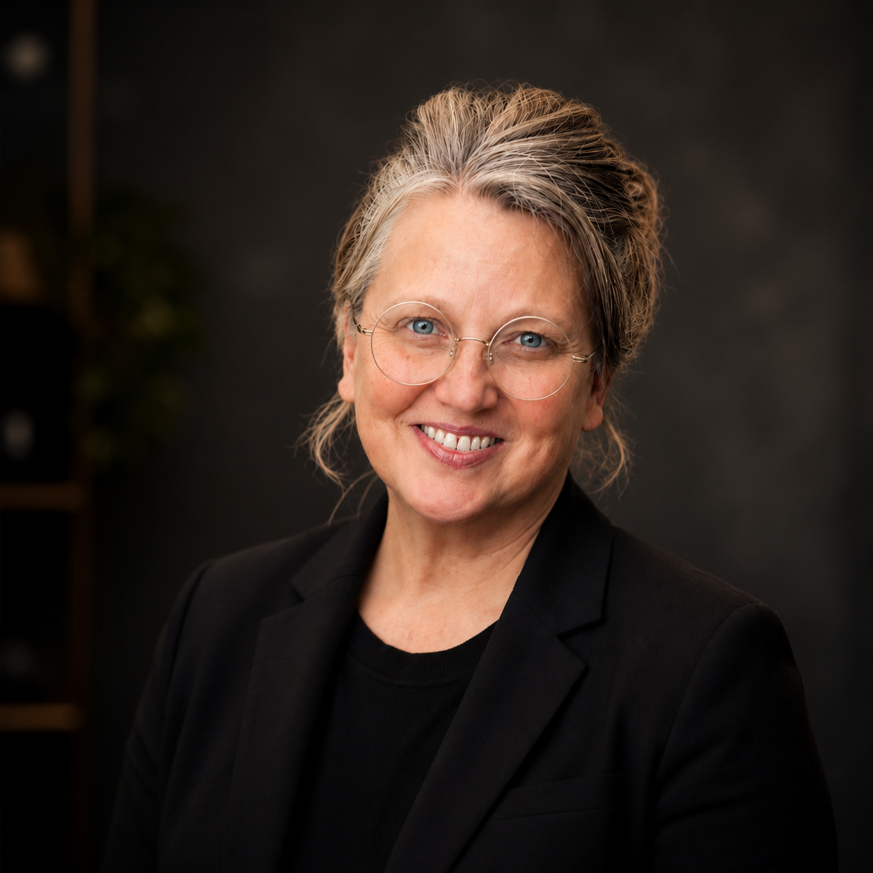 Portrait of a smiling woman with short gray hair, glasses, wearing a black blazer, against a dark background.