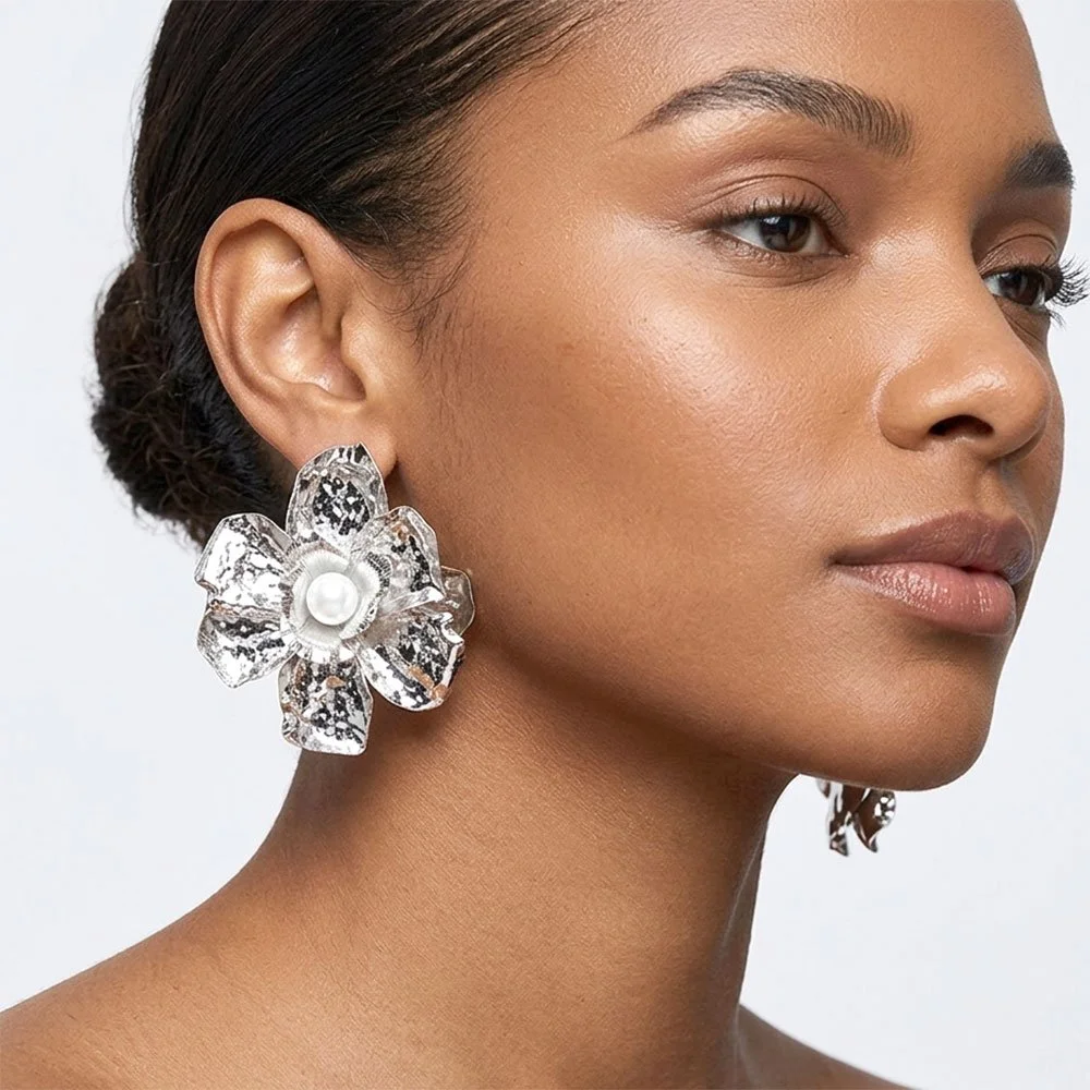 Close-up of a woman's face with tan skin and clear skin, wearing large silver floral earrings with pearl centers, short dark hair, and neutral makeup.