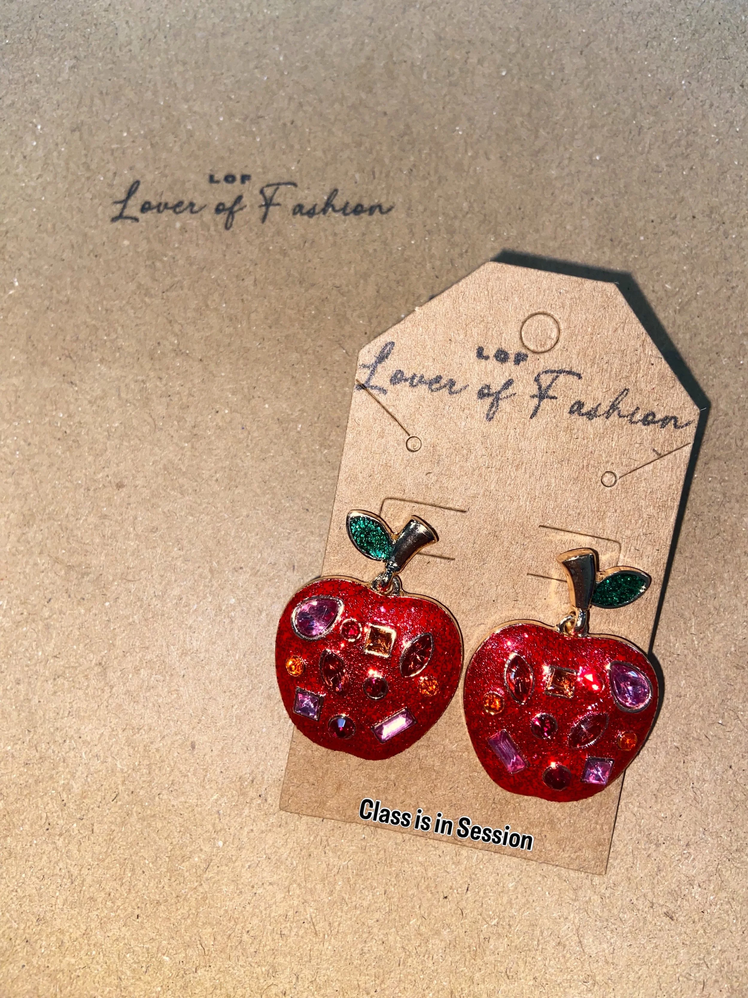 Red shiny apple-shaped earrings with green leaves, attached to a cardboard backing with the text 'Lover of Fashion' written twice and an additional note 'Class is in Session' at the bottom.