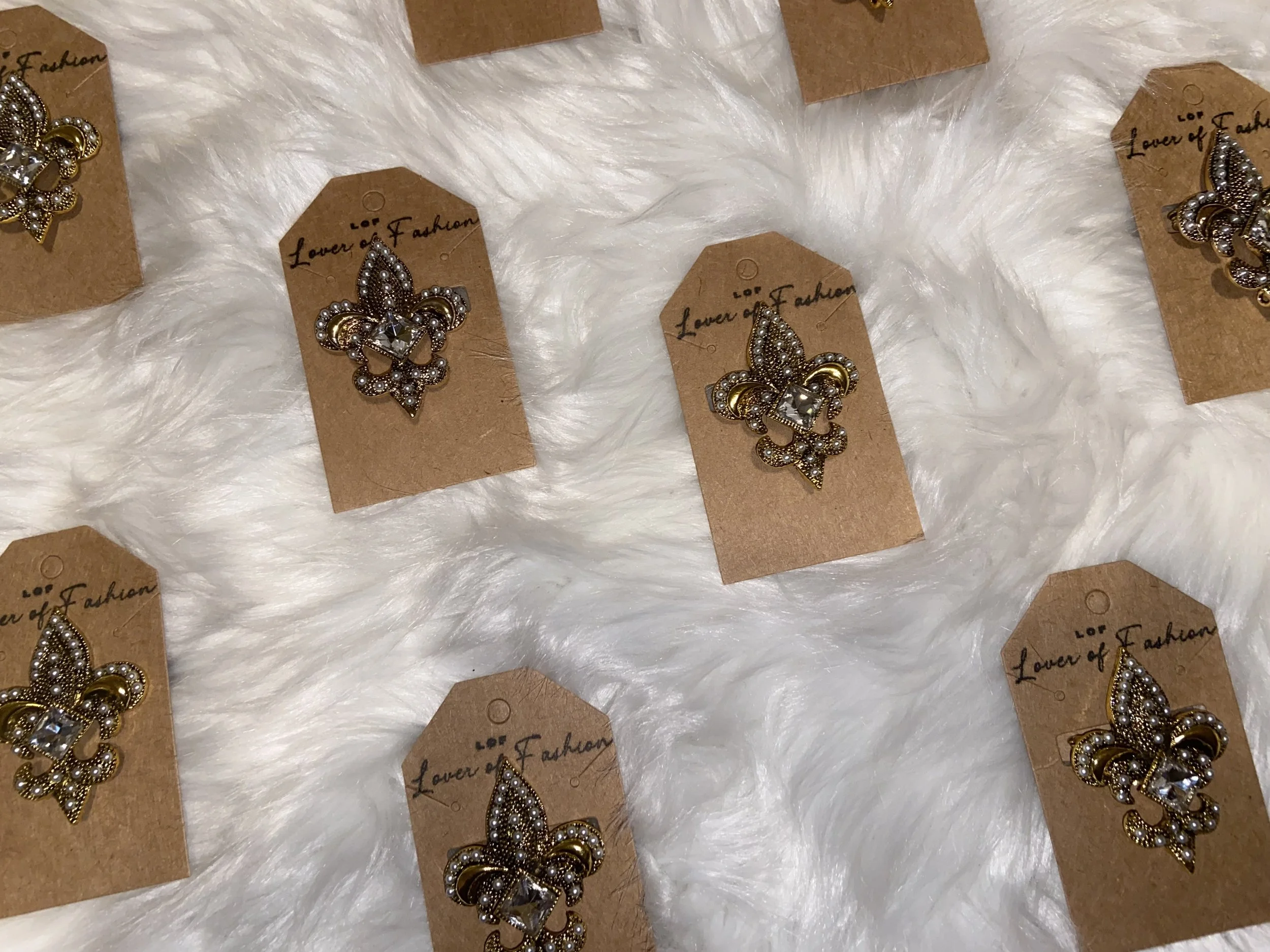 A collection of decorative jewelry clips with an anchor or Fleur de Lis design, attached to brown tags that read "Lover of Fashion," displayed on a white furry surface.