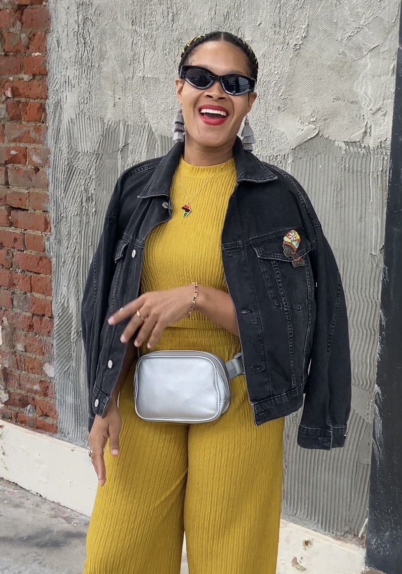 A woman wearing sunglasses, a black denim jacket, a yellow pleated outfit, and jewelry, standing against a concrete wall with a brick wall to her side, smiling and holding her hand near a silver fanny pack.