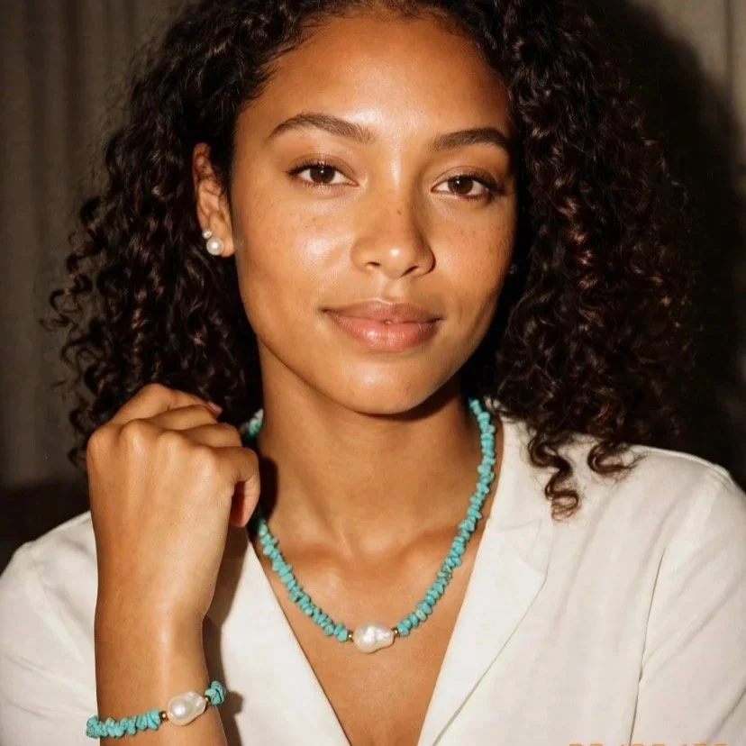 A woman with curly hair, wearing pearl earrings, a turquoise necklace with a large pearl, and a matching bracelet with a pearl. She is dressed in a white top and has a confident, relaxed expression.