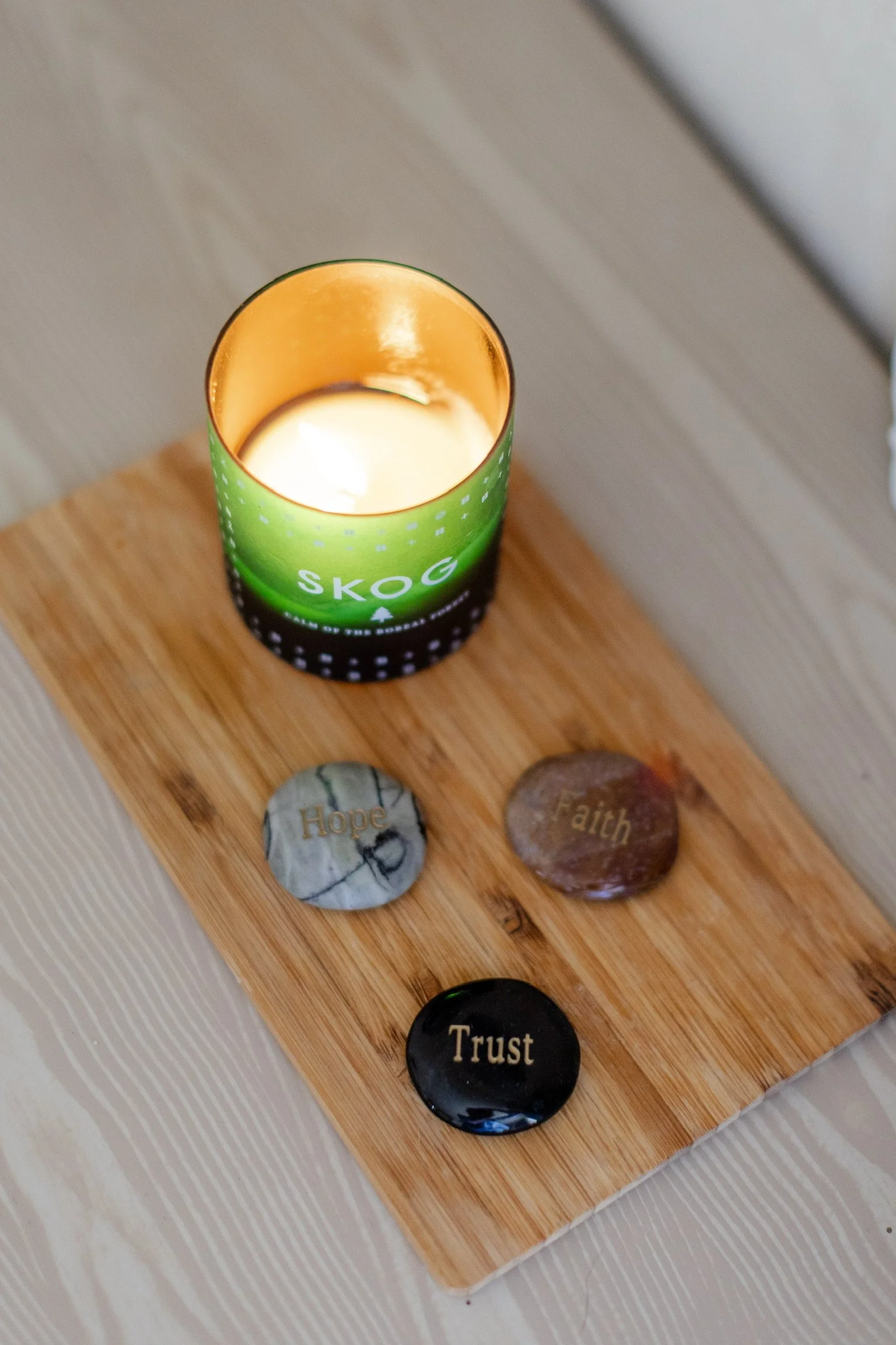 A wooden board with five decorative stones with inspirational words placed on it, and a green candle holder labeled 'Skog' with a lit candle inside.