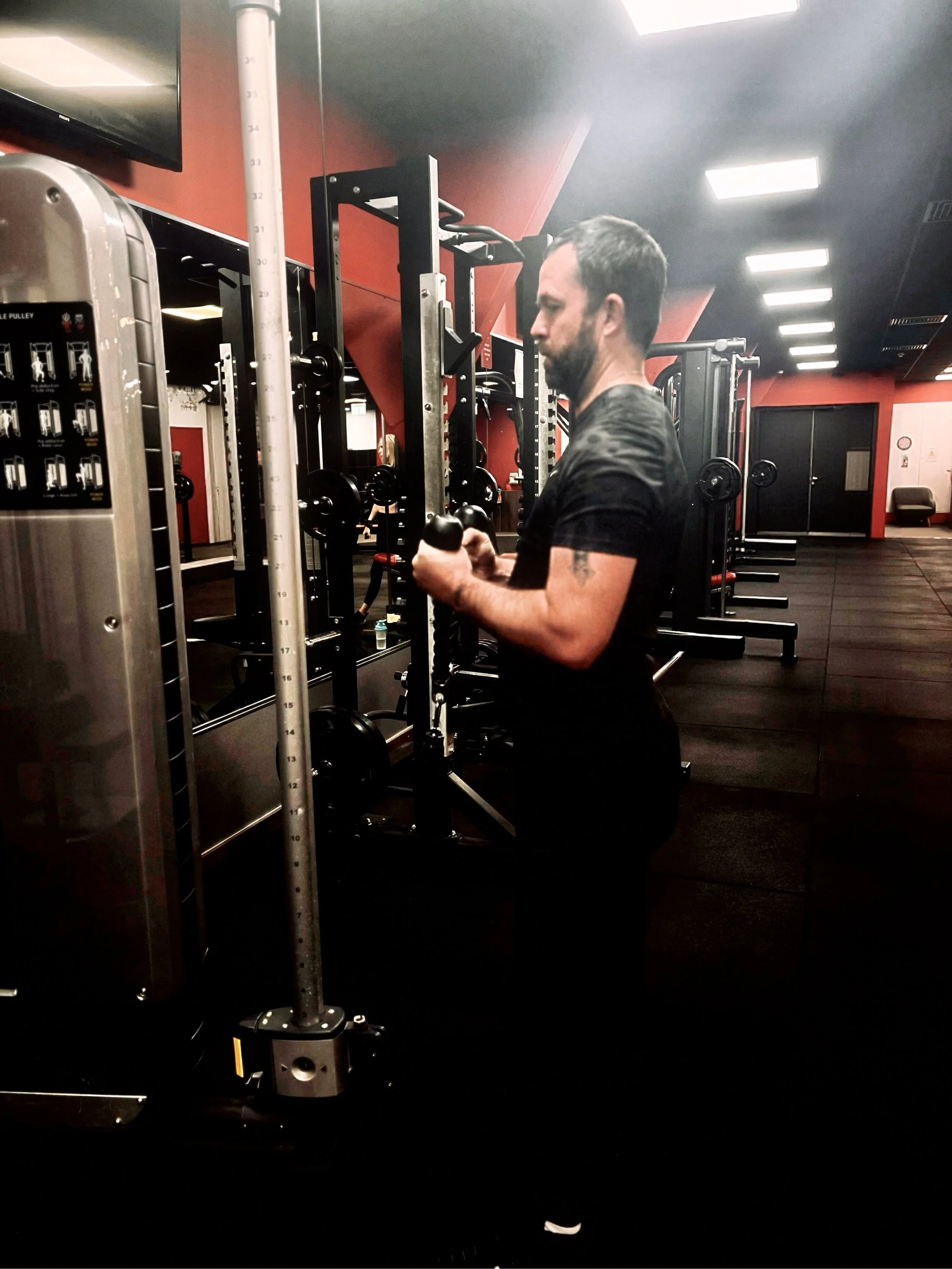 A man with dark hair and a beard in a dark gym is standing next to gym equipment, holding a black kettlebell. The gym has black floors and red walls with overhead bright lights.