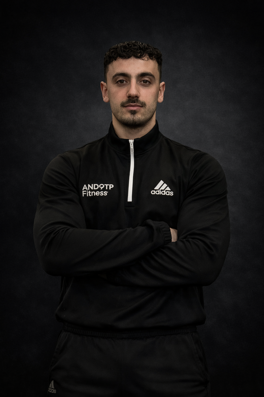 A young man in a black Adidas sports jacket with arms crossed, standing against a dark background.