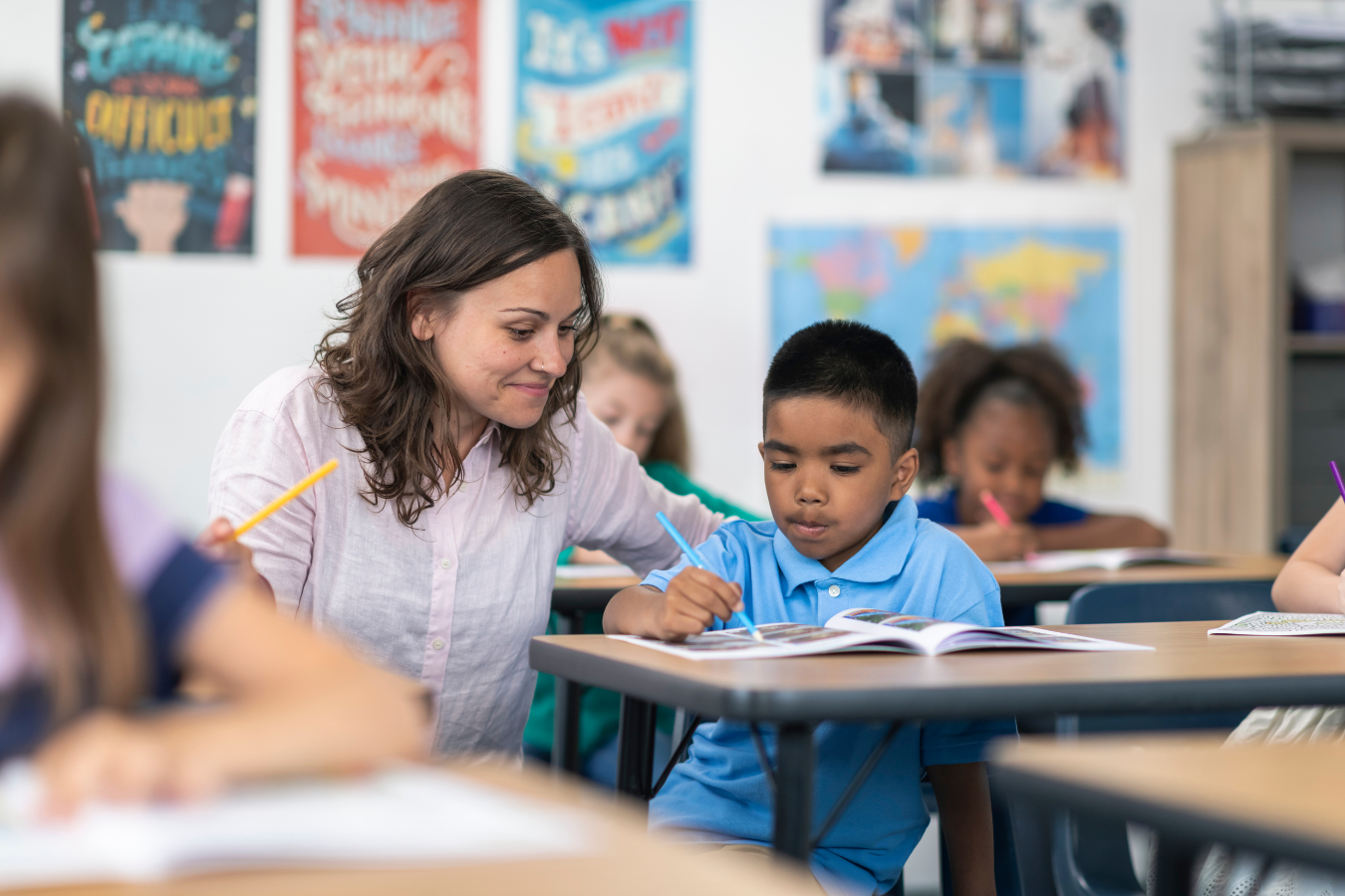 A classroom scene with a teacher assisting a young student at his desk. The teacher is smiling and holding a yellow pencil, while the student is focused on his open book, as other students work in the background.