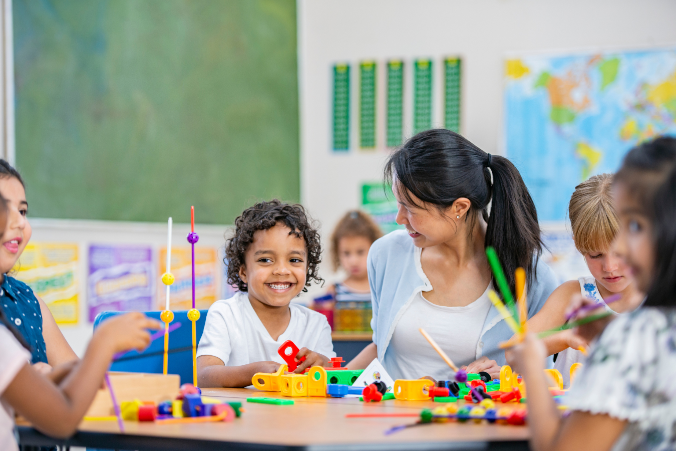 Teacher smiling and interacting with children playing with colorful educational toys in a classroom.