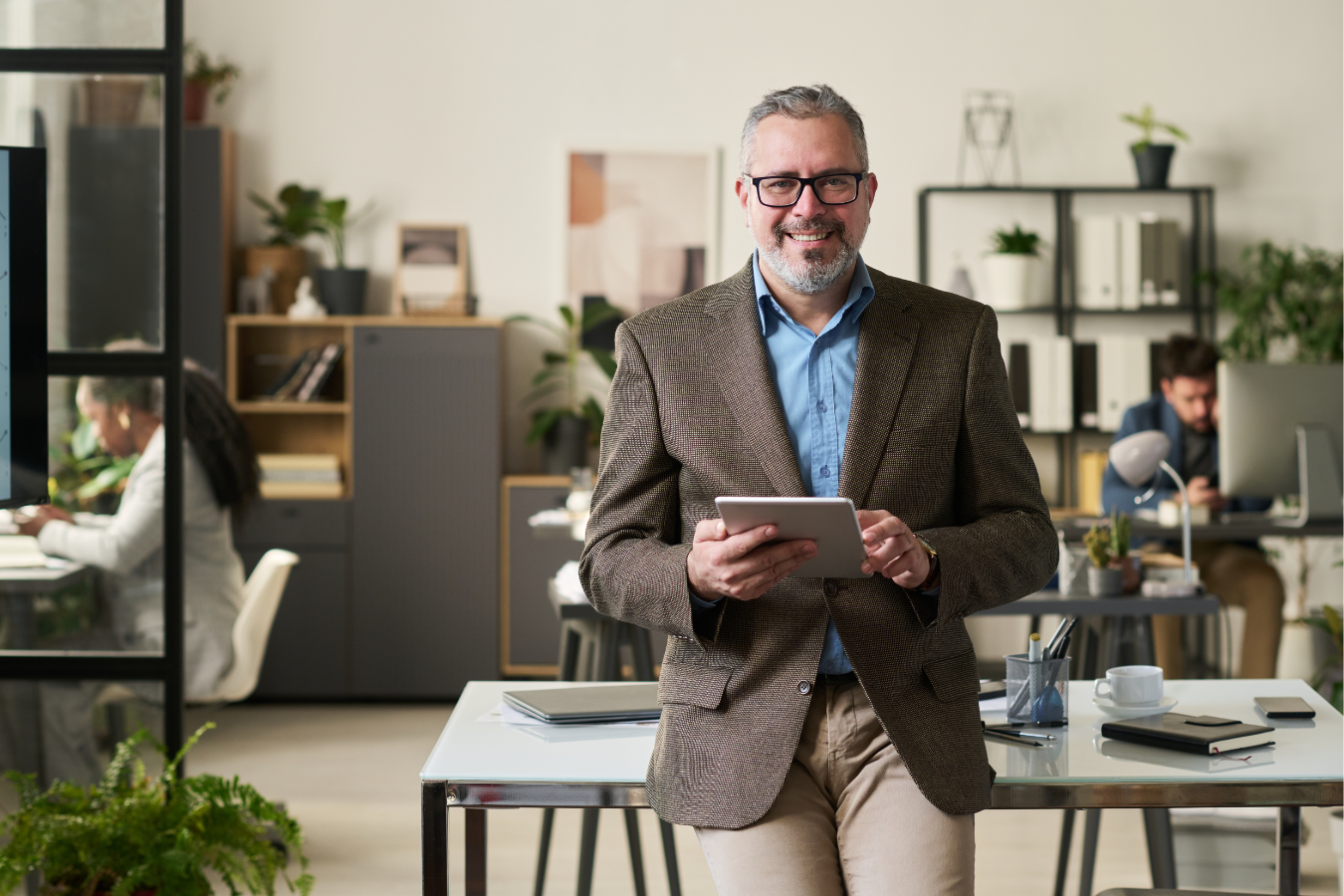 A middle-aged man with glasses, gray hair, and a beard, wearing a brown blazer and blue shirt, smiling and holding a tablet in a modern office with two other professionals working in the background.