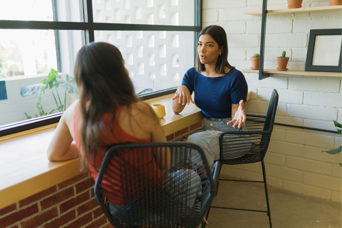 Two women having a conversation in a cafe, one wearing a blue top and the other in a red top, sitting by a large window with potted plants on the windowsill.