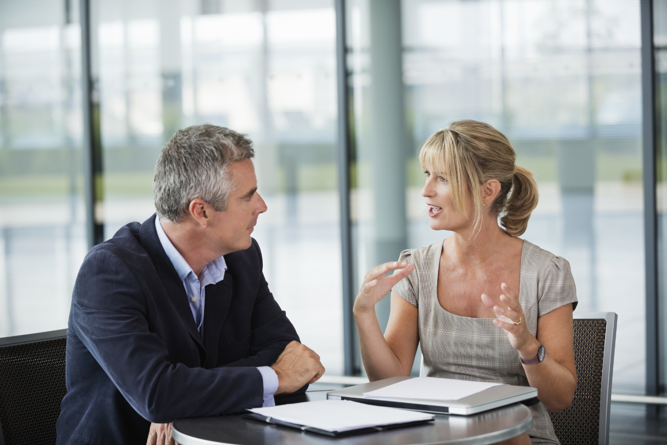 A man and woman having a conversation at a table in a modern office with large windows.