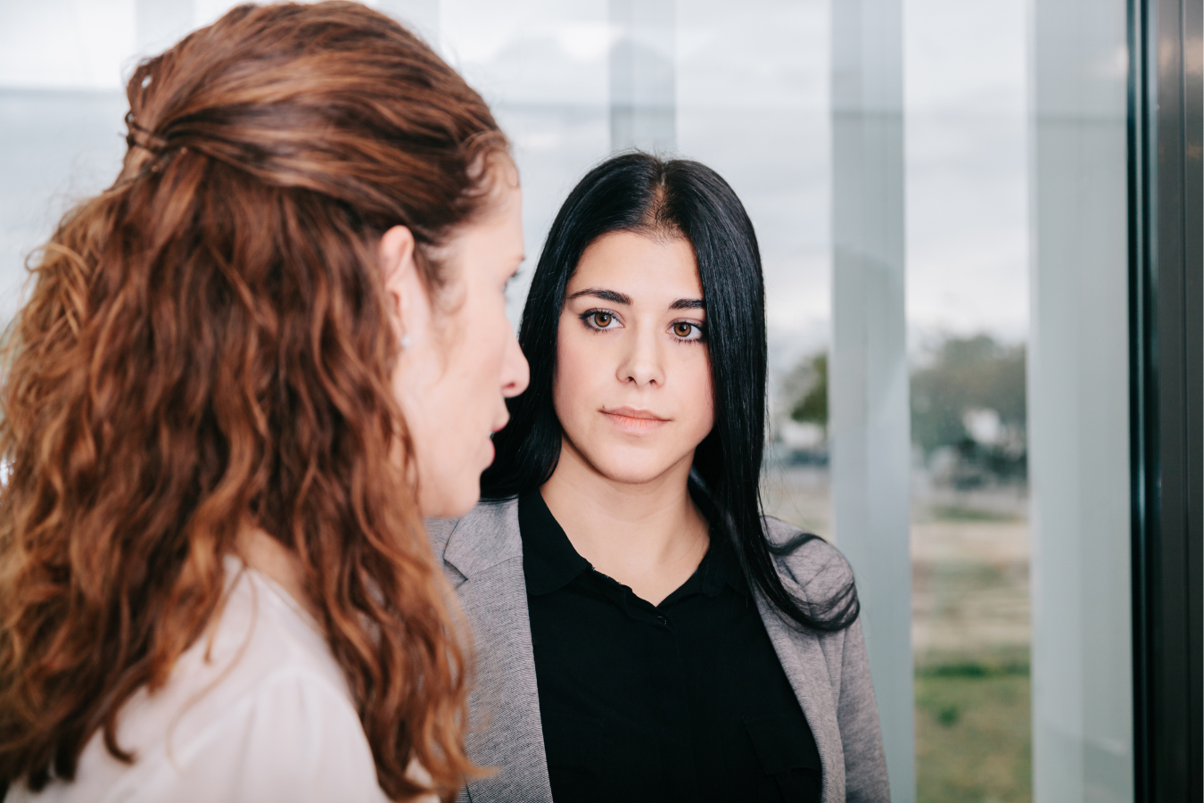Two women having a serious conversation in an office building, with one woman with long red hair and the other with straight black hair.