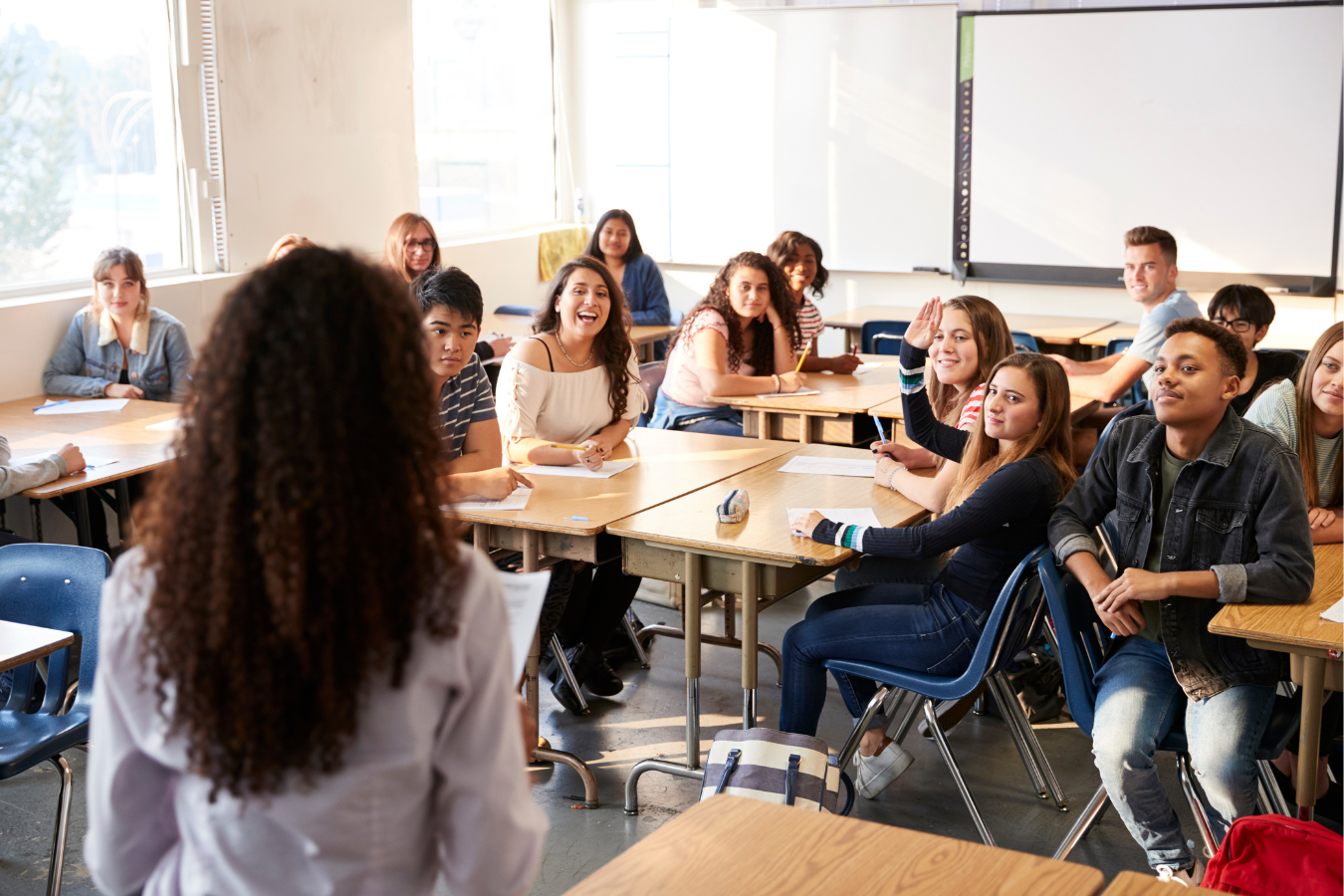 Students in a classroom, some raising hands, listening to a teacher.