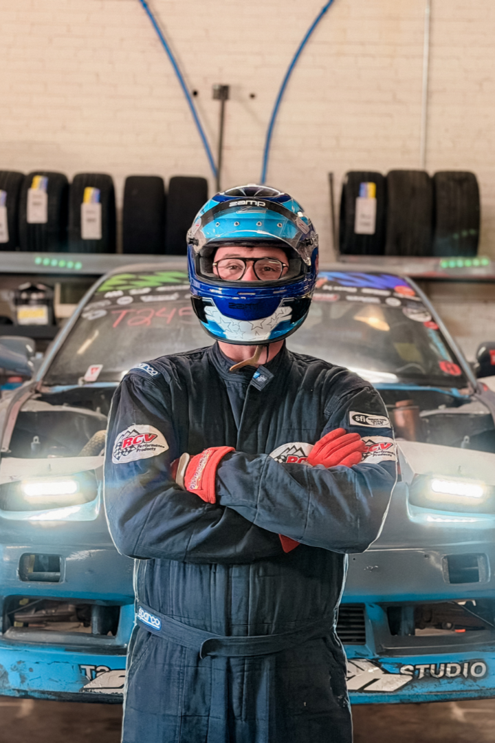 Racing driver in racing suit and helmet standing with arms crossed in front of a race car inside a garage or workshop. Mr. Machine racing episode.