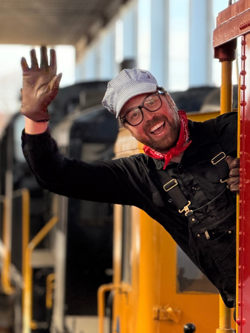 A man dressed as a train conductor, wearing a striped hat, black outfit, glasses, and a red bandana, is smiling and waving from the window of a train car. Mr. Machine train episode.