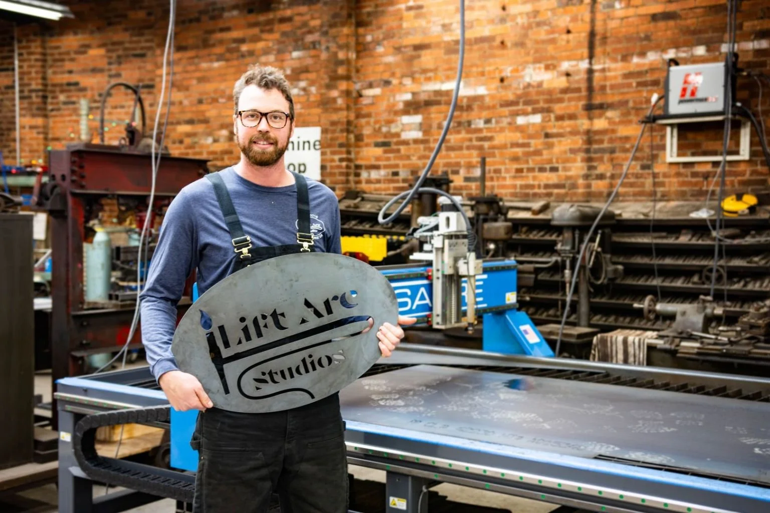 A man with glasses and a beard smiling, holding a large metal sign that reads 'Lift Arc Studios' in a workshop with tools, machinery, and industrial equipment in the background. Tay Whiteside.