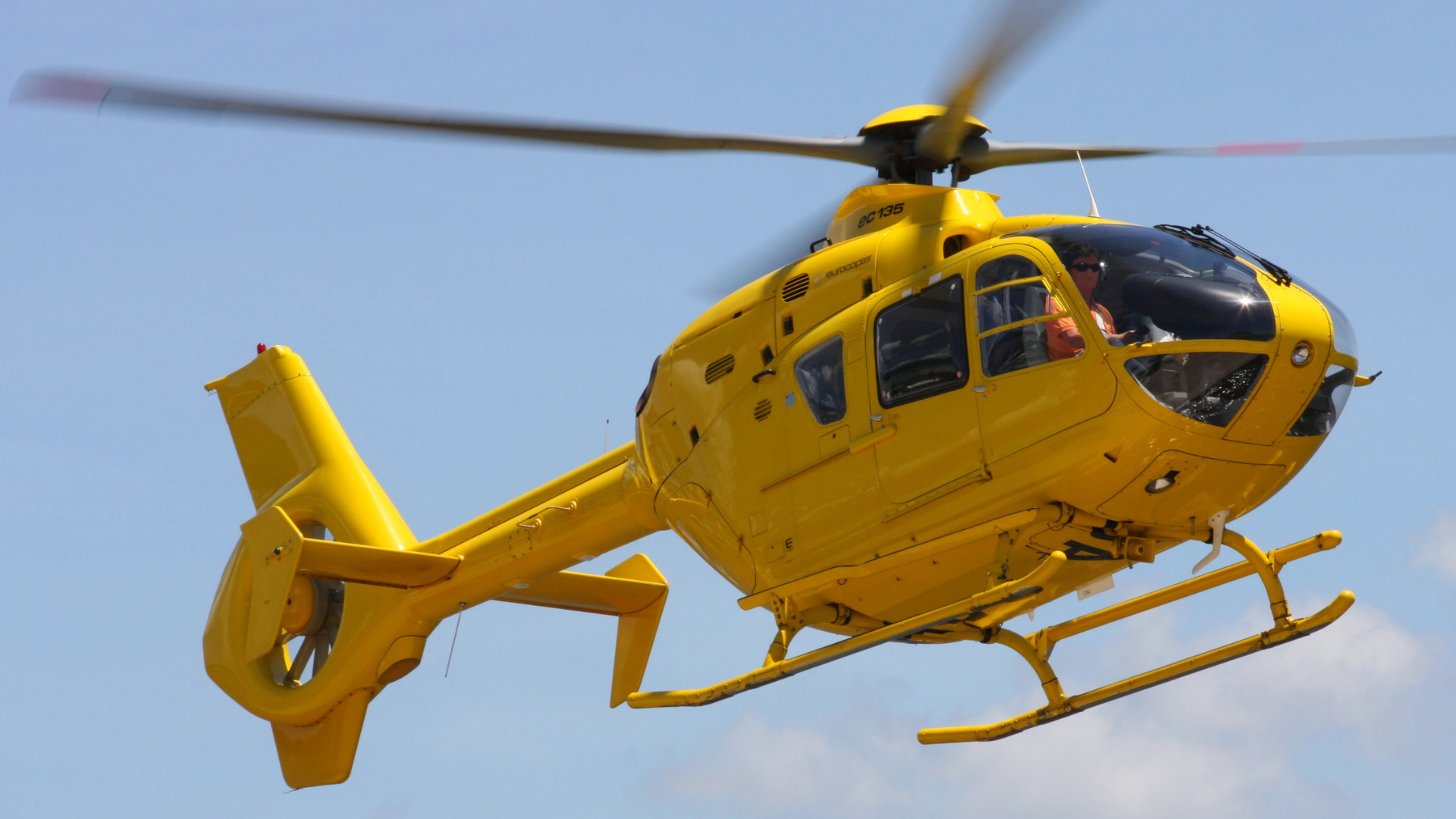 A yellow helicopter flying in the sky with a clear blue background, pilot visible through the cockpit window. Helicopters for kids. Helicopter show for kids. 