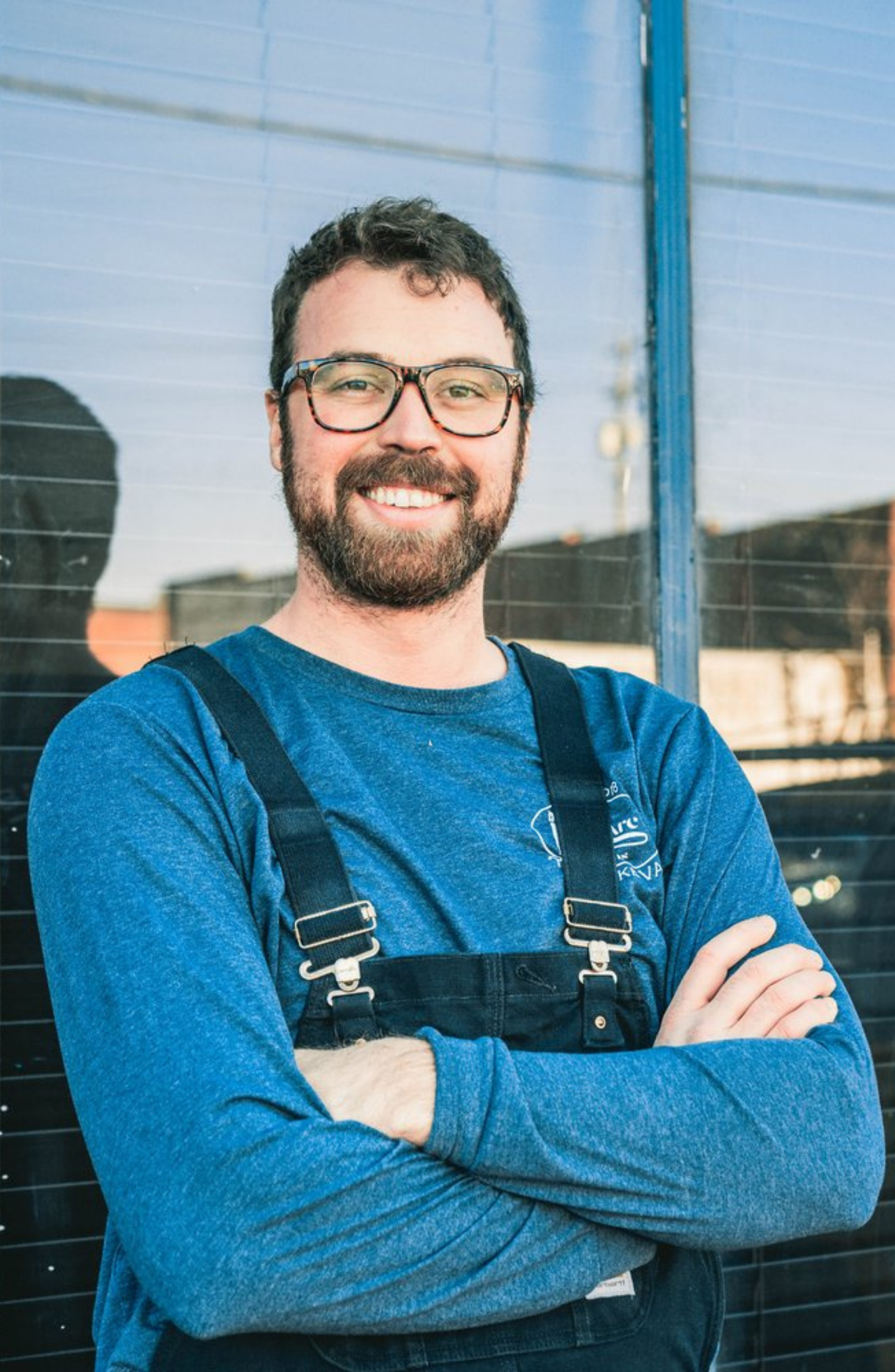 A man with glasses, a beard, and curly hair smiling with arms crossed standing in front of a reflective surface with a building and sky in the background. Tay Whiteside from Lift Arc Studios.