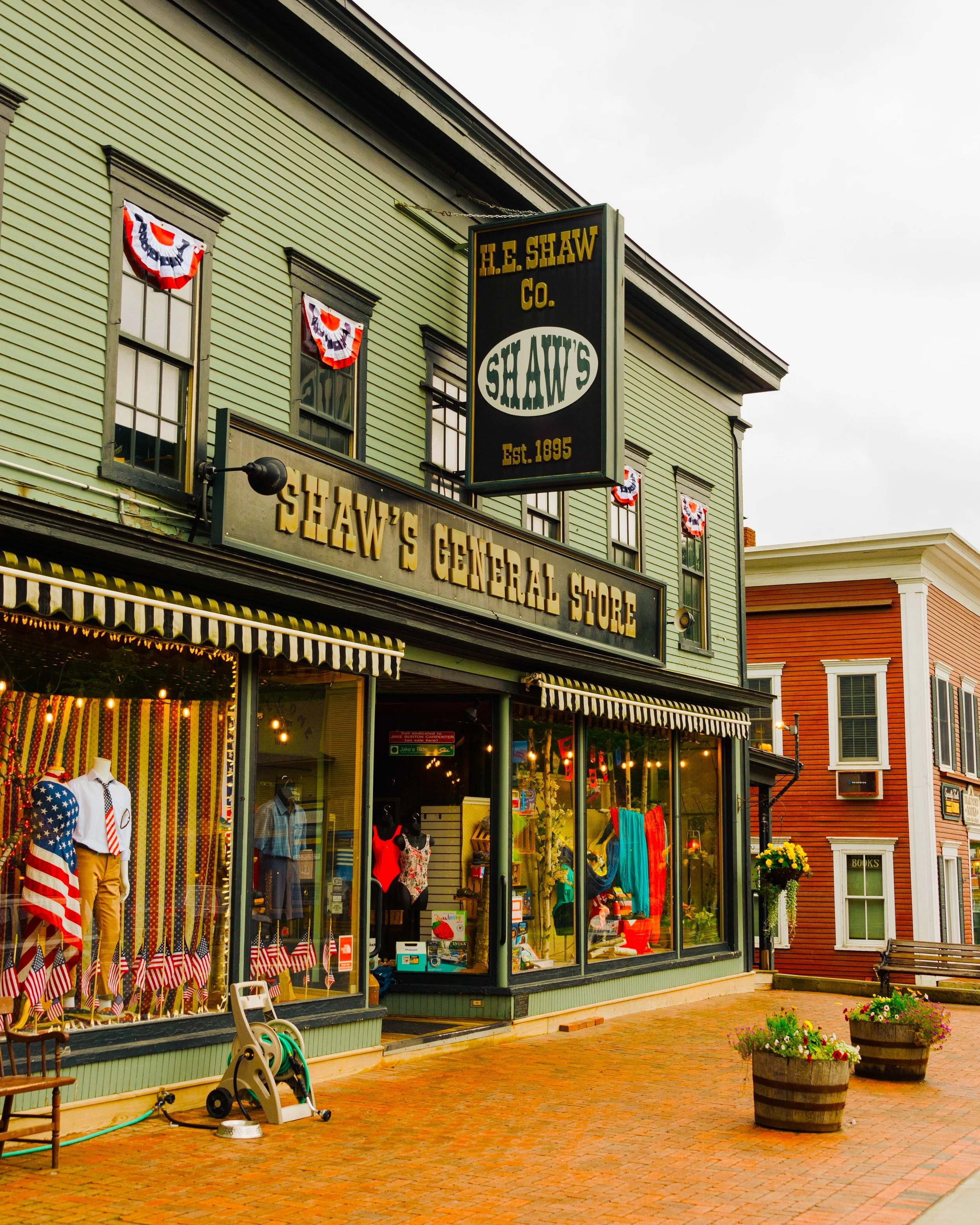 Street view of Shaw's General Store with patriotic decorations in the windows. Storefront has large glass windows displaying clothing and household items. Two large flower pots with colorful flowers are outside on a brick walkway. The building has a 
