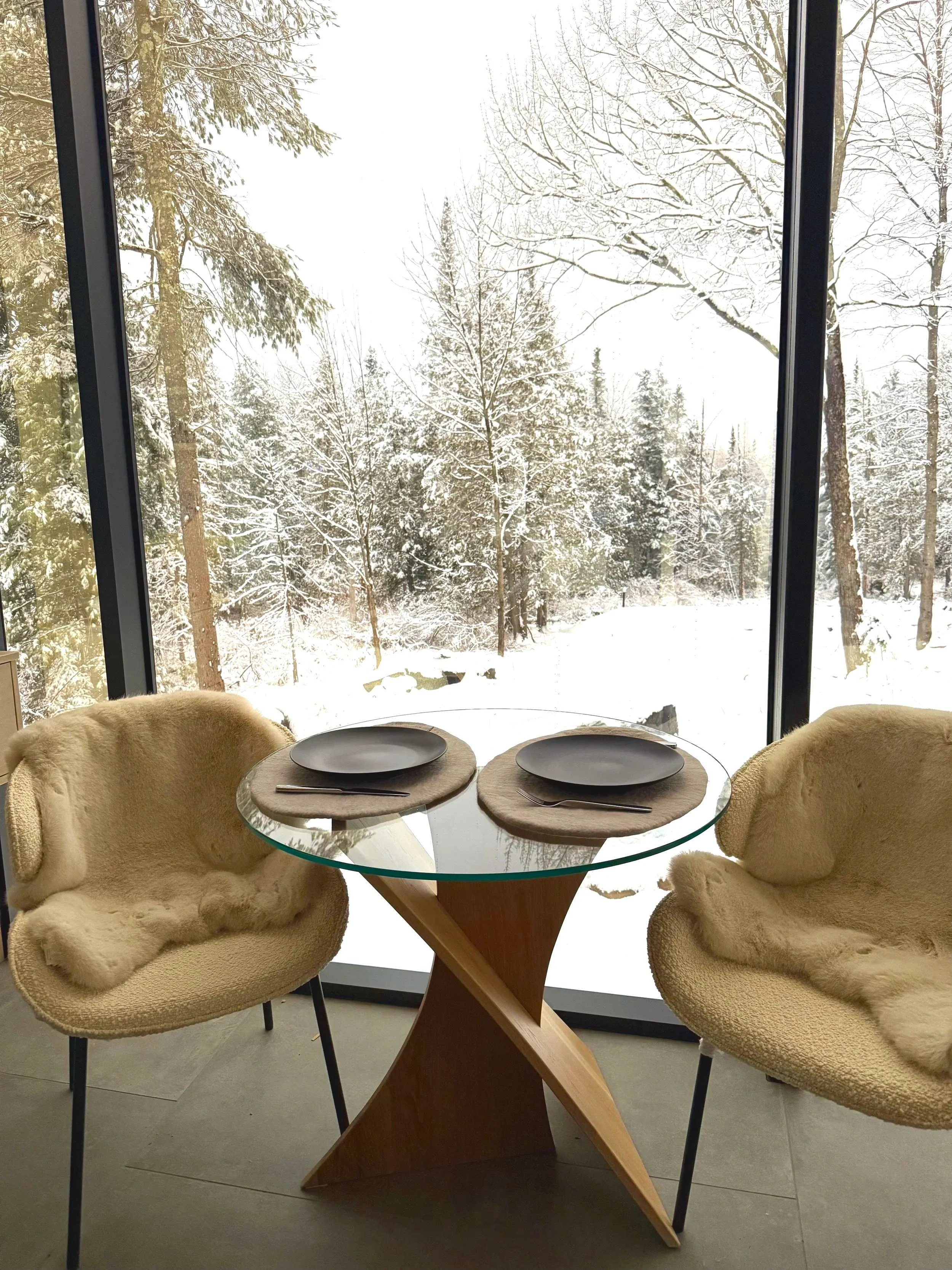 A round glass dining table set with two placemats, plates, and silverware, with two plush beige armchairs on either side, near large floor-to-ceiling windows showing a snowy winter landscape with trees covered in snow.