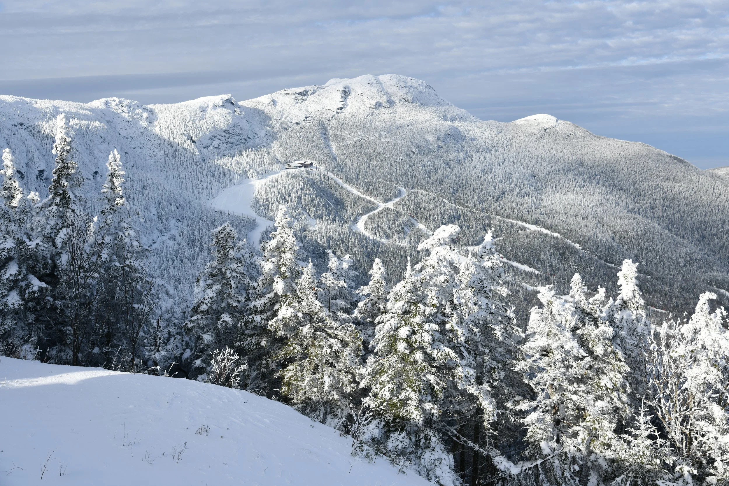 Snow-covered mountain landscape with trees, a building, and winding paths under a partly cloudy sky.