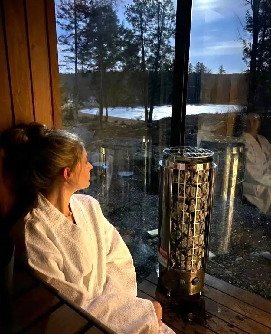 A woman sitting in a sauna or bathhouse wearing a white towel, looking out a large window at a snowy outdoor landscape with trees, with a heater for sauna rocks in front of her.