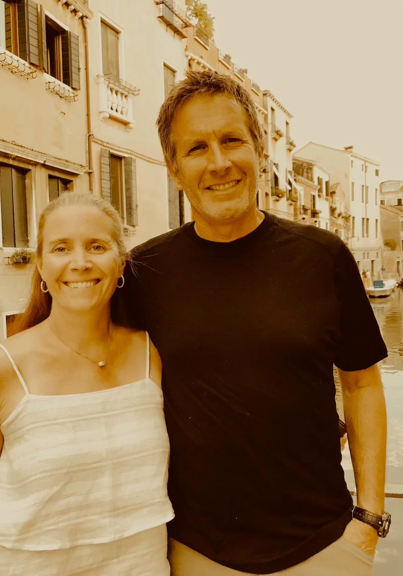 A man and woman standing outdoors with a canal and European-style buildings in the background, during sunset or golden hour.