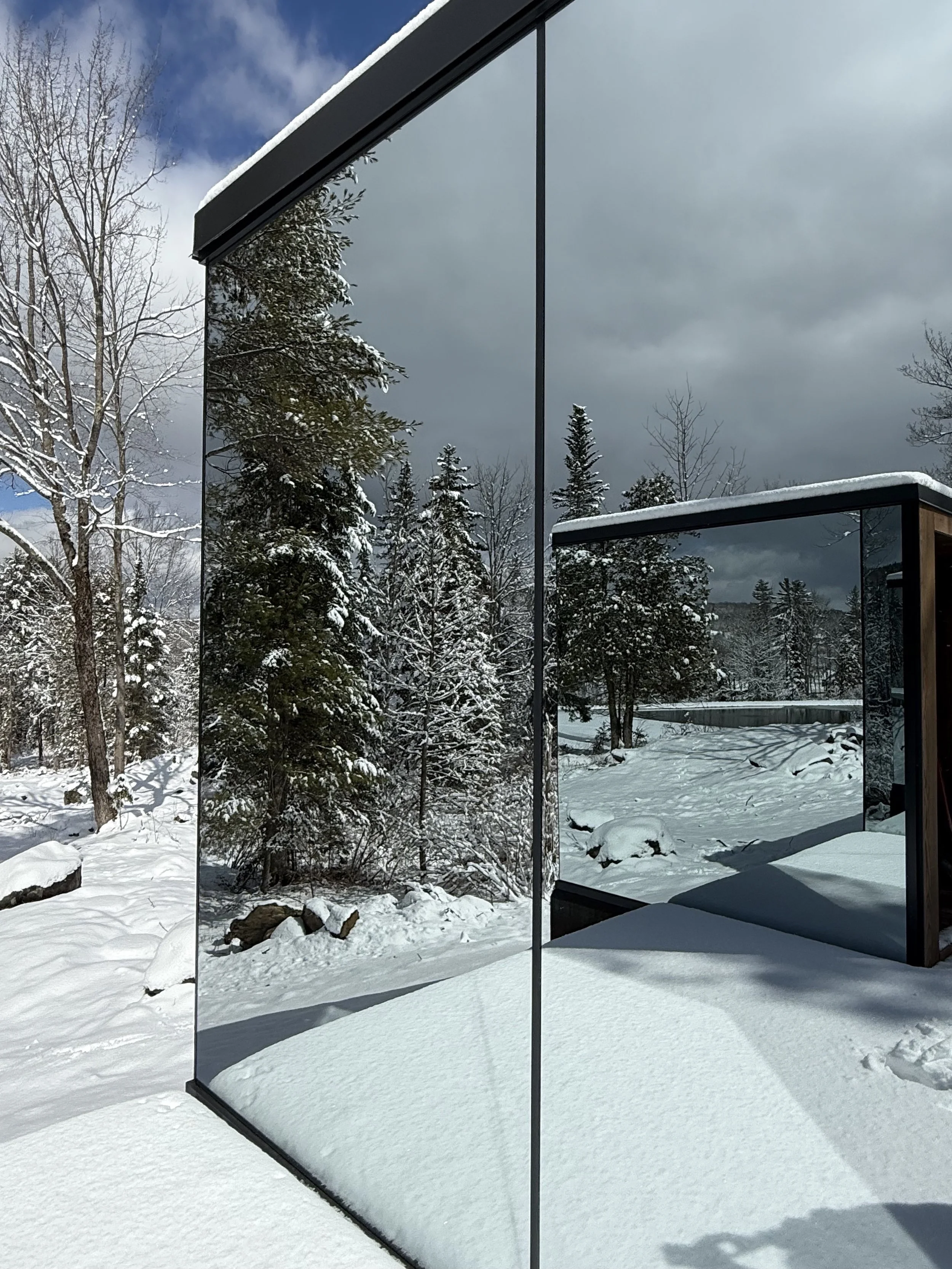 Snow-covered ground and trees reflected in a large outdoor glass mirror with a dark frame, mounted on a stand, in a wintry landscape with cloudy sky.
