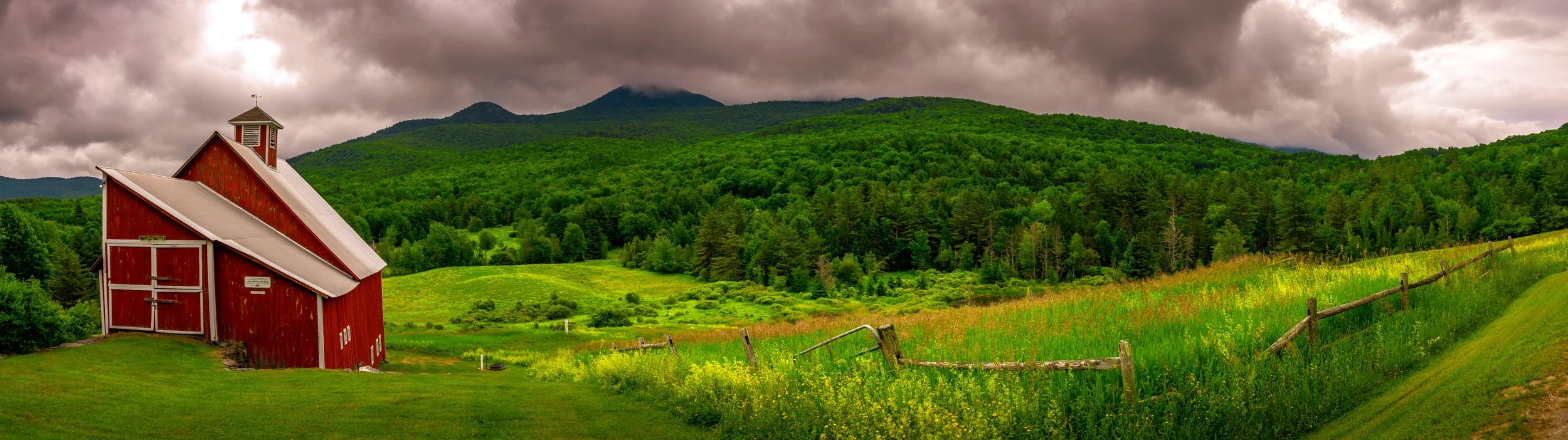 A red barn on a lush green hillside with mountains and dark, cloudy sky in the background.