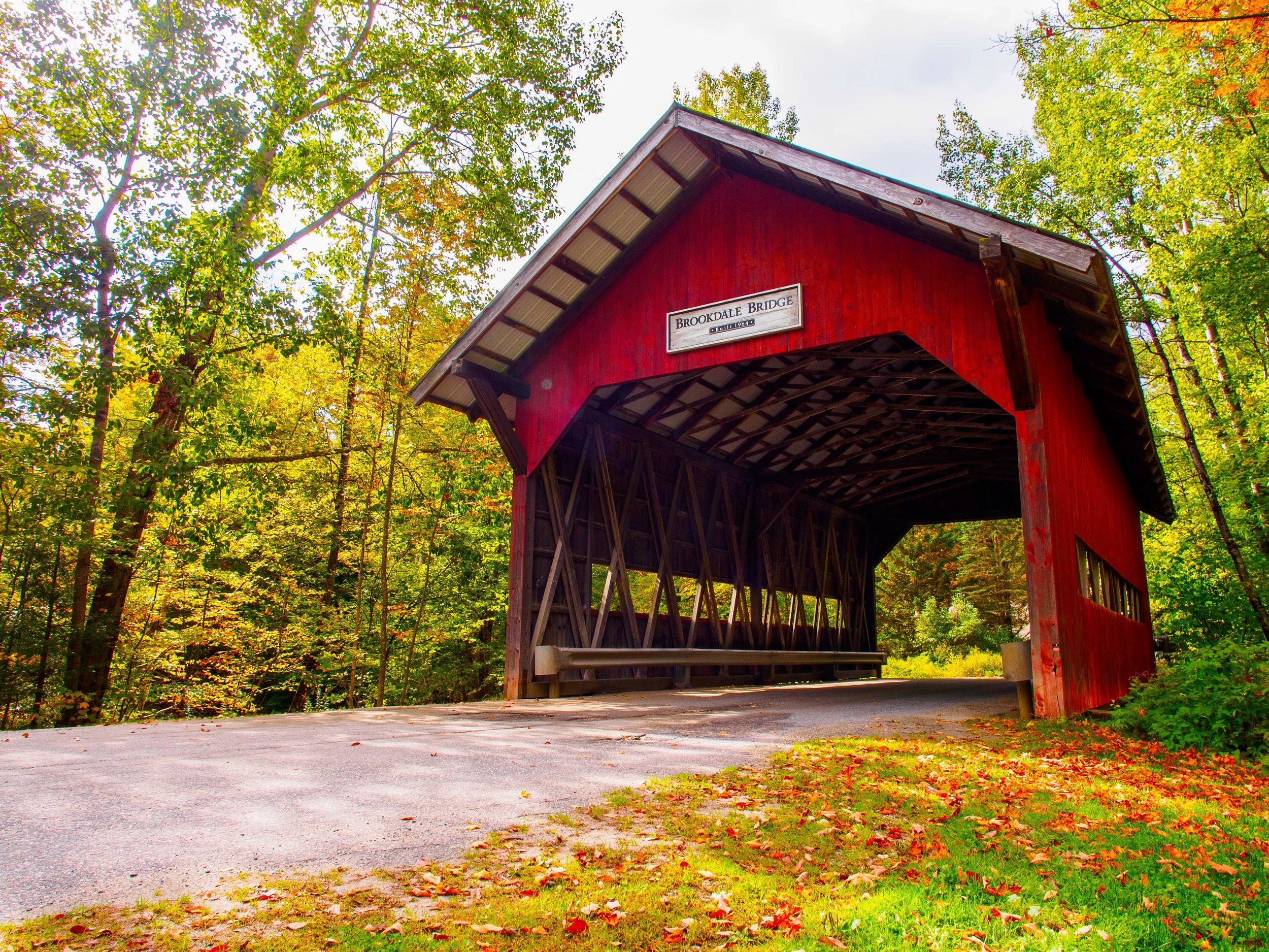 A red covered bridge named Brodke Bridge, built in 1964, surrounded by trees with green and autumn-colored leaves, during daytime with an overcast sky.