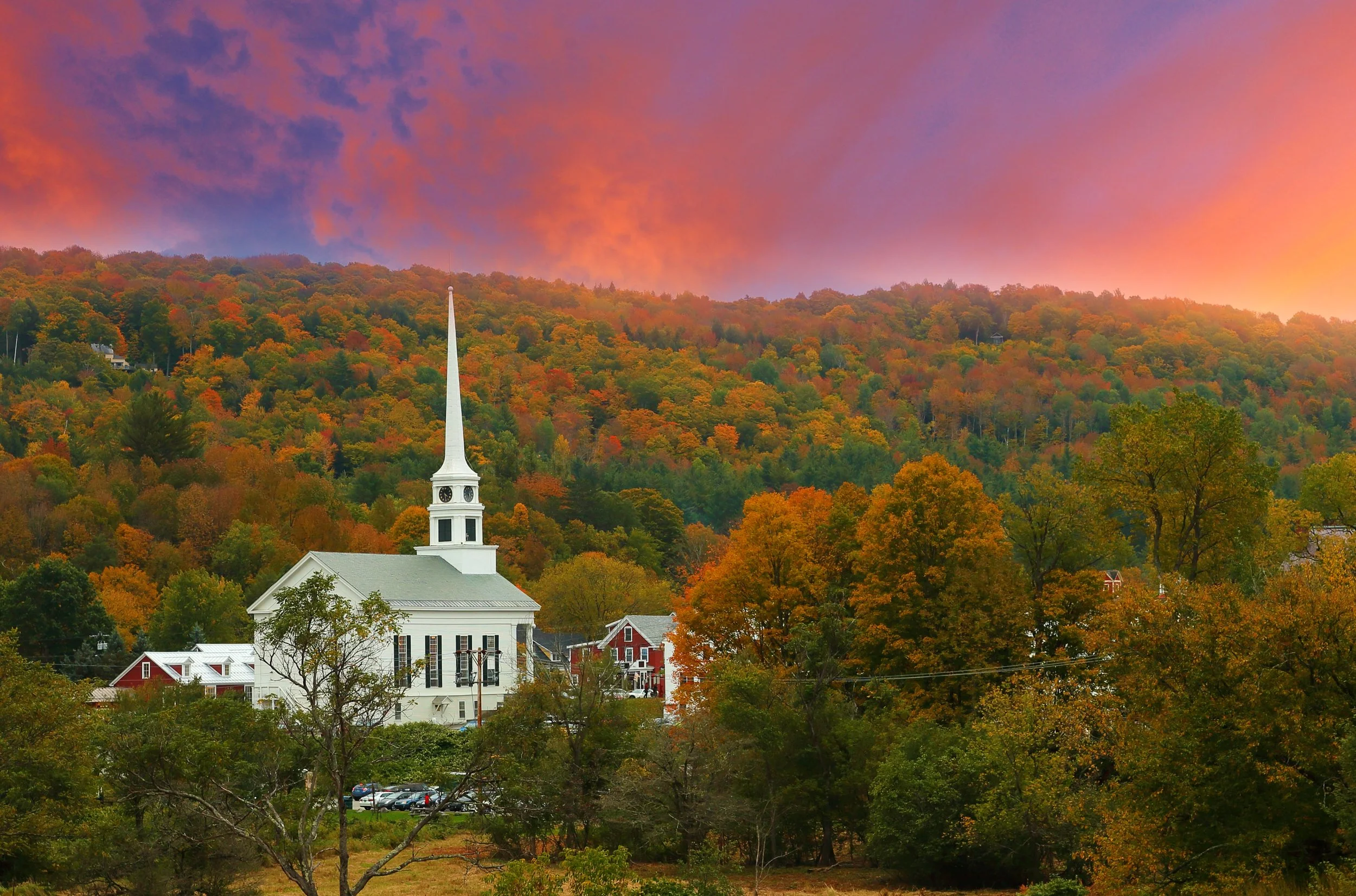 A small town with a white church surrounded by autumn trees and colorful fall foliage, under a vibrant sunset sky filled with pink, purple, and orange clouds.
