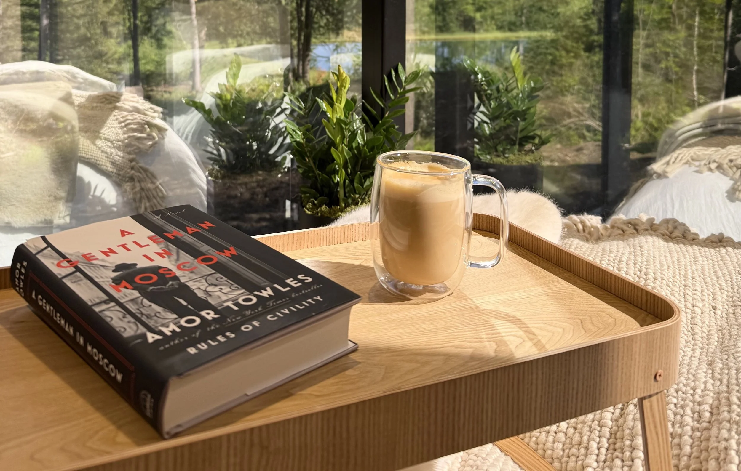 A wooden tray on a table holding a book titled 'A Gentlemen in Moscow' and a glass mug of coffee or latte. The background shows a large window with green plants and outdoors scenery.