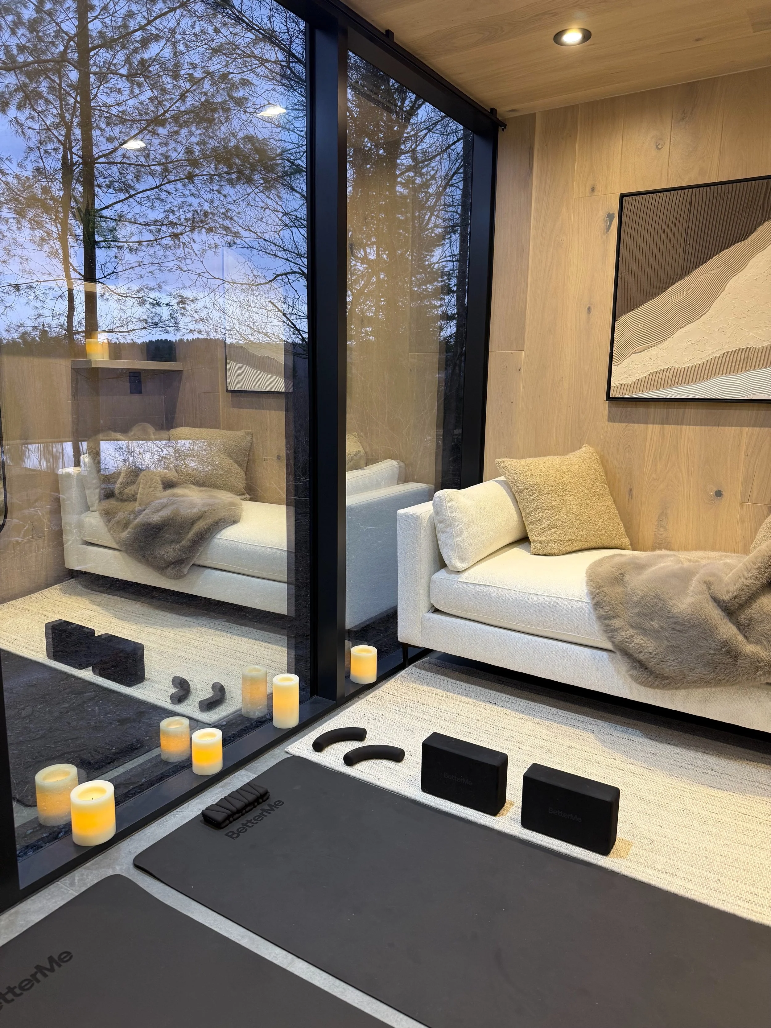 Interior of a cozy room seen through a glass wall, with two white sofas, a wooden wall with a minimalistic painting, candles, workout mats, and fitness equipment for home exercise.