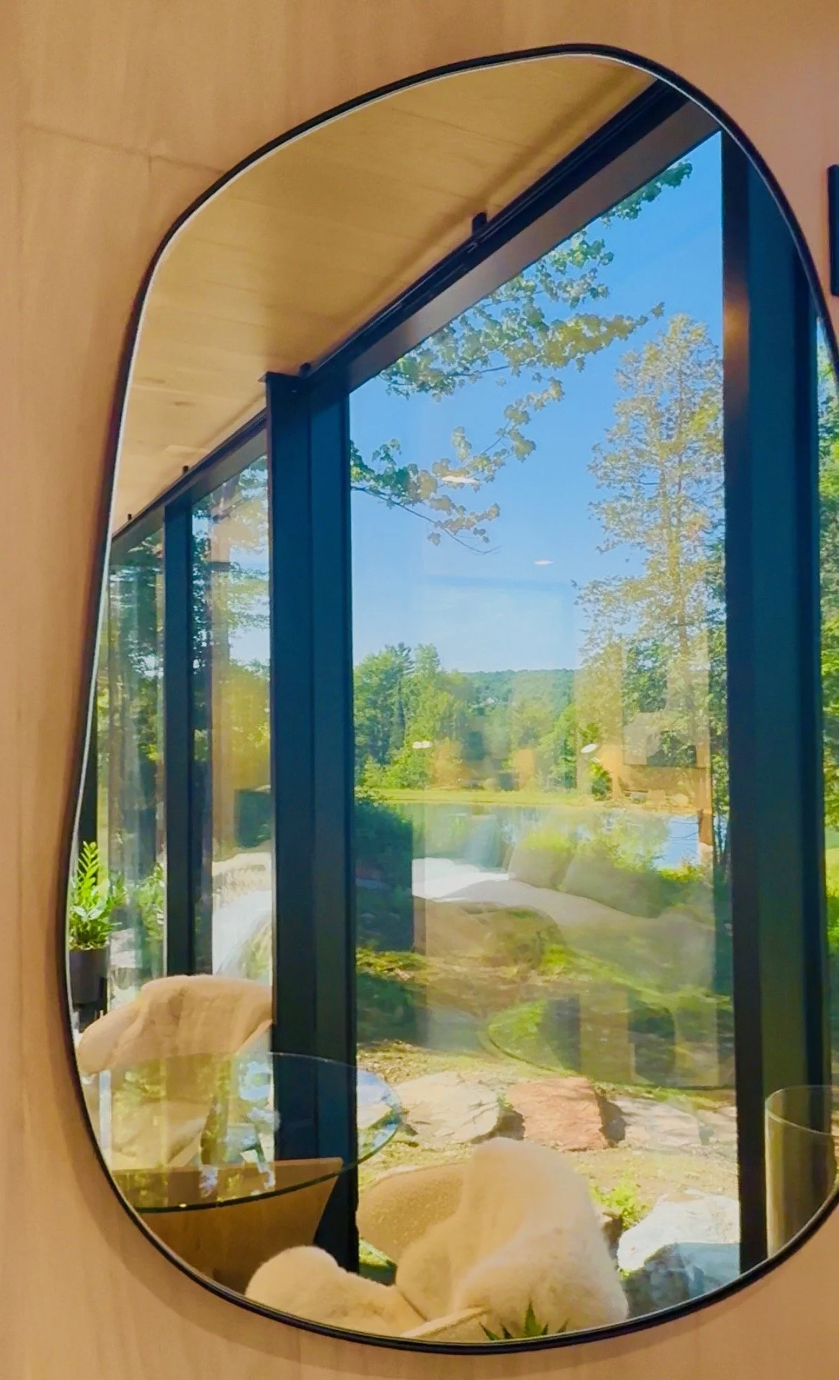 A mirror with a black frame reflecting a landscape scene with trees, a pond, blue sky, and a pathway.