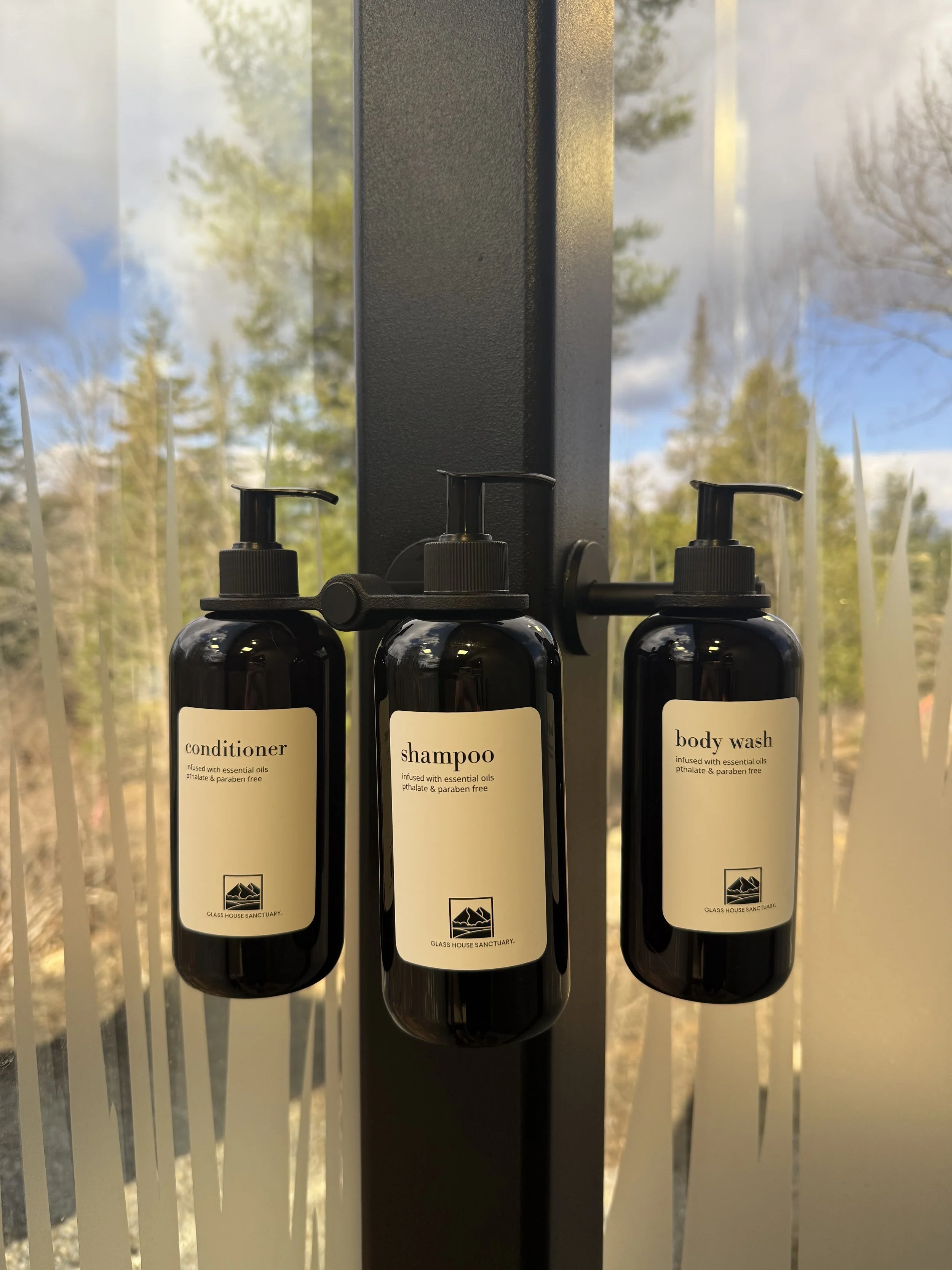Three black bottles labeled conditioner, shampoo, and body wash hang from hooks against a window with blinds and a background of trees and sky.