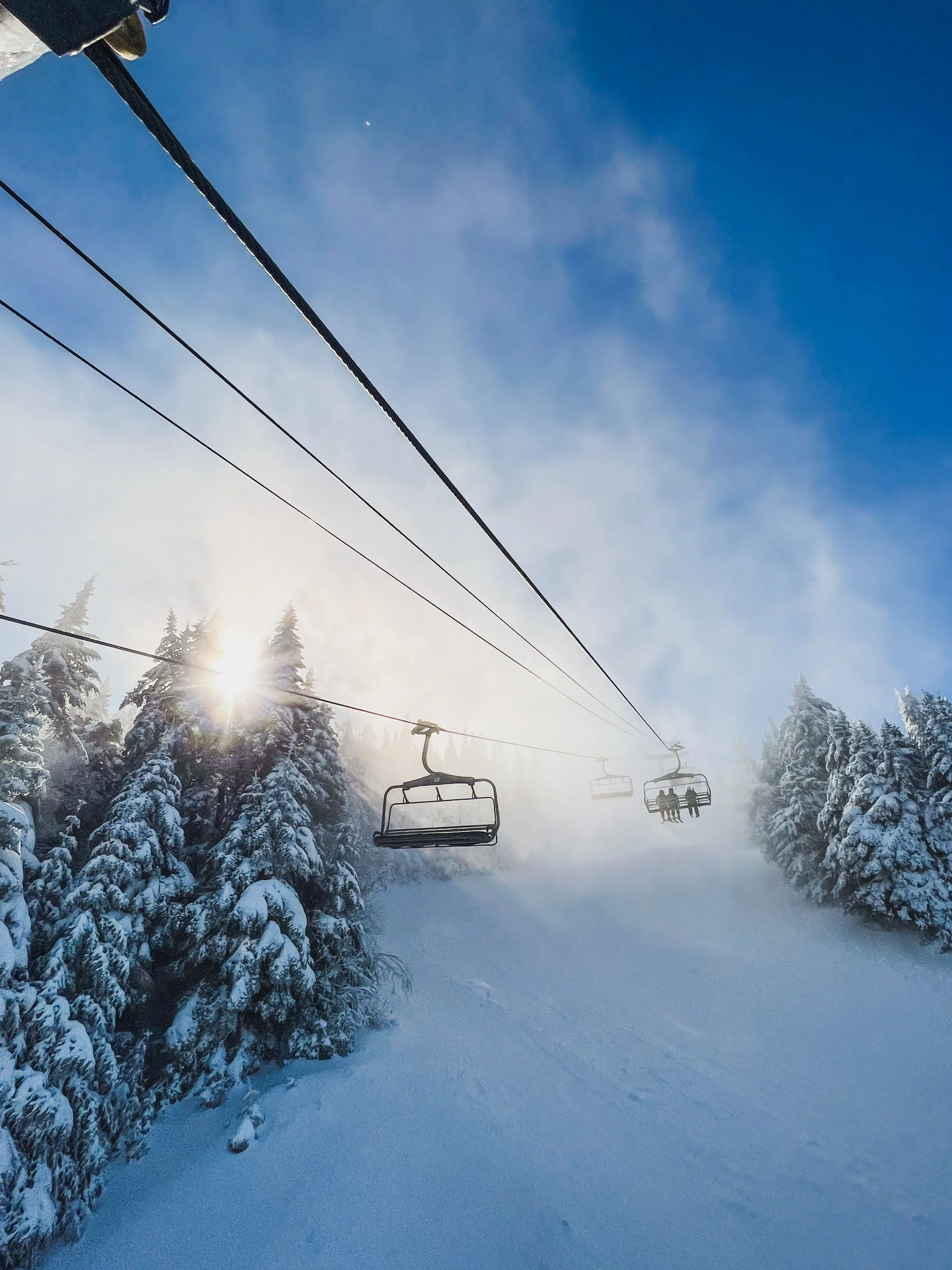 Ski lift carrying two people over a snow-covered forest with a clear blue sky and the sun shining through the trees.